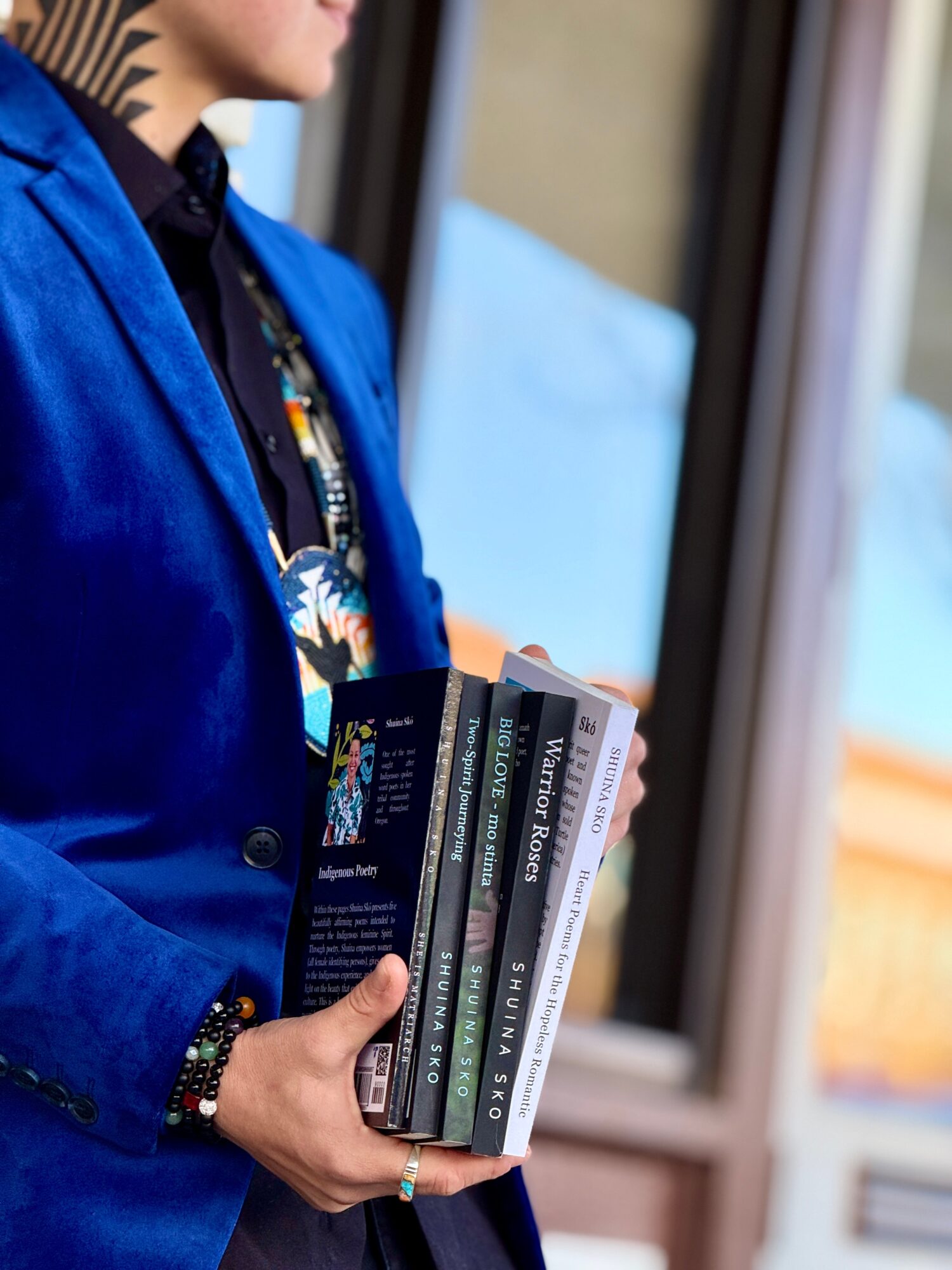 Person holding a stack of books near a window, wearing a blue jacket and patterned shirt.