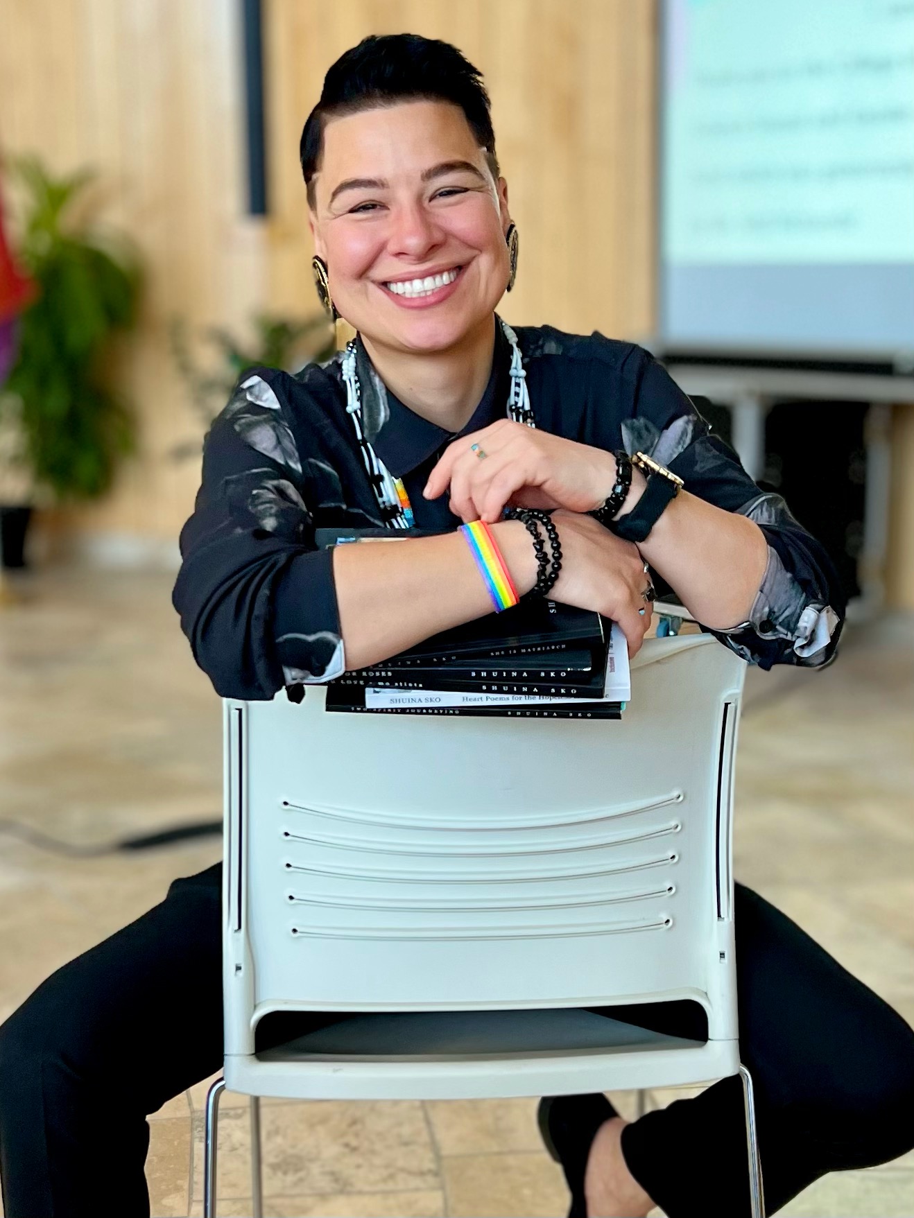 Person smiling, sitting on the floor with arms resting on a chair, in a classroom setting with a screen in the background.
