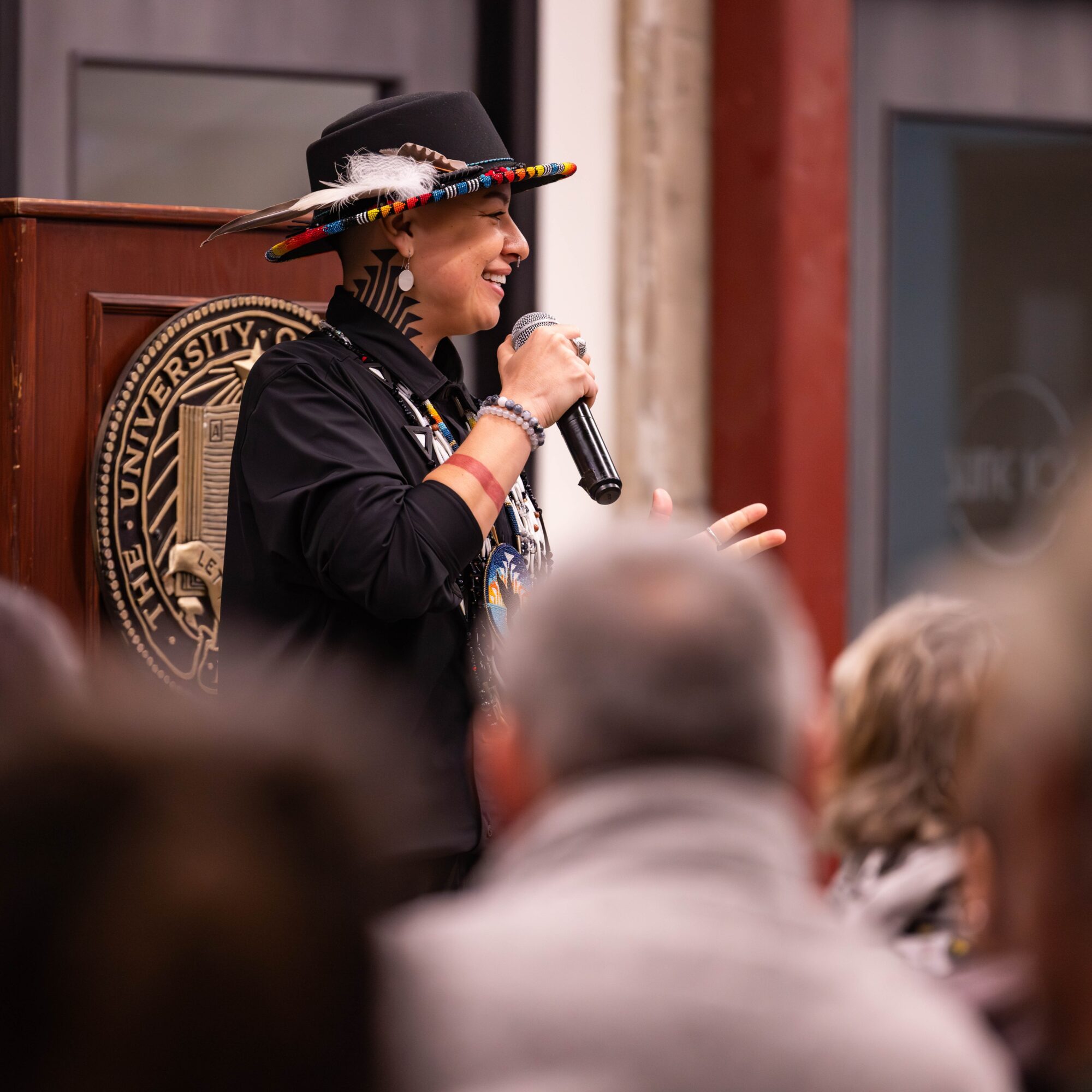 Woman speaking into microphone at event, wearing a colorful hat and black outfit, with audience in foreground.
