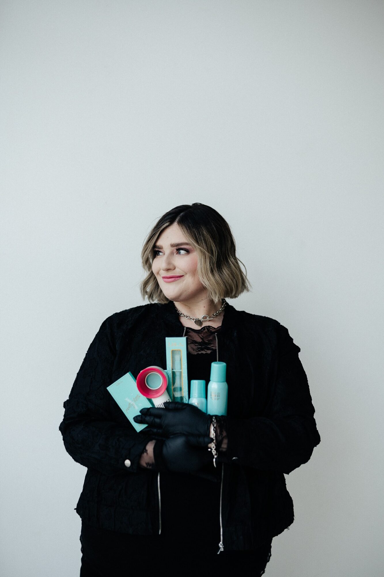 Woman with shoulder-length wavy hair holding skincare products, wearing a black jacket and gloves, standing against a plain background.
