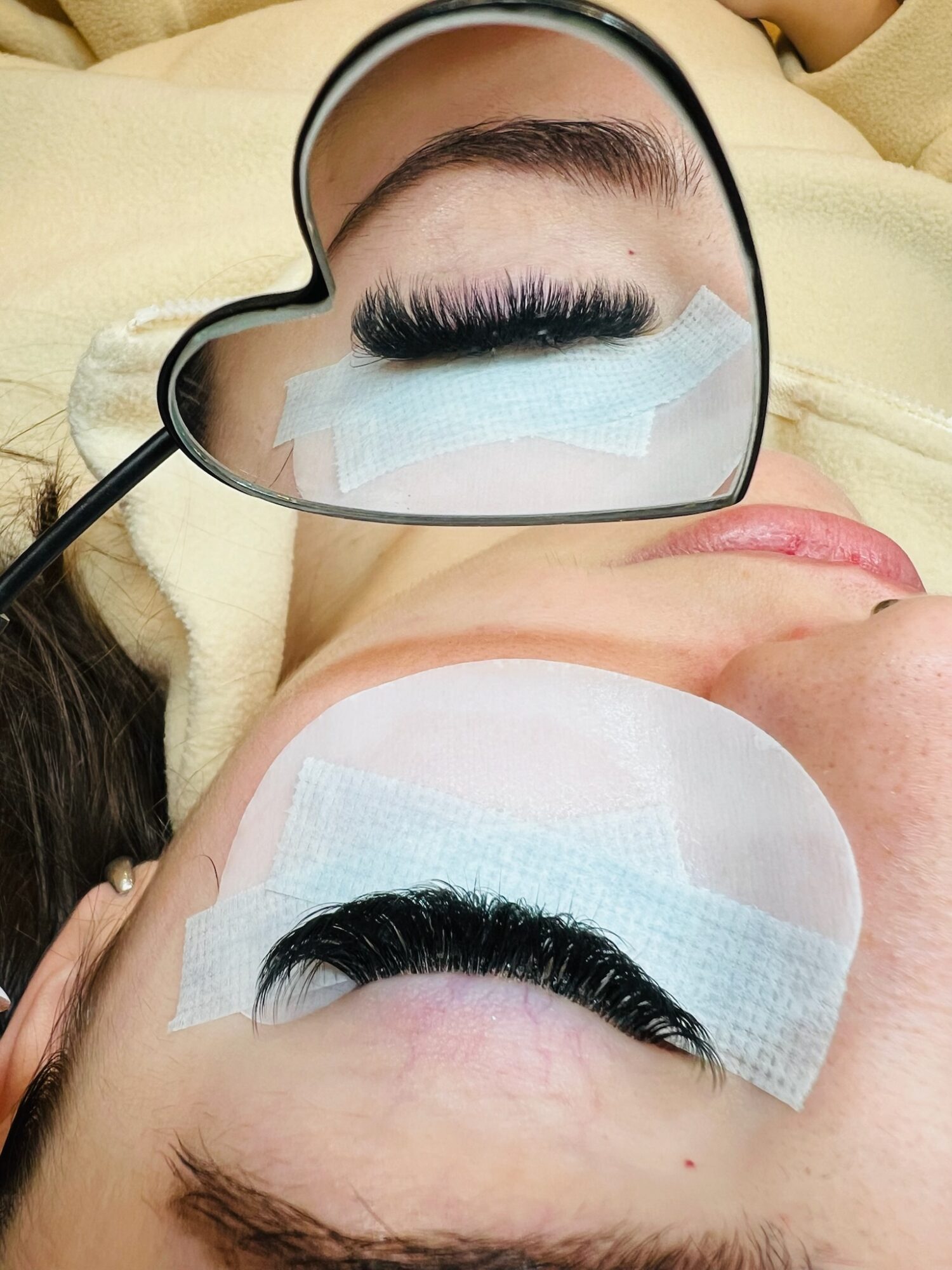 Two eyes with eyelash extensions, one reflected in a heart-shaped mirror, with protective tape on eyelids.