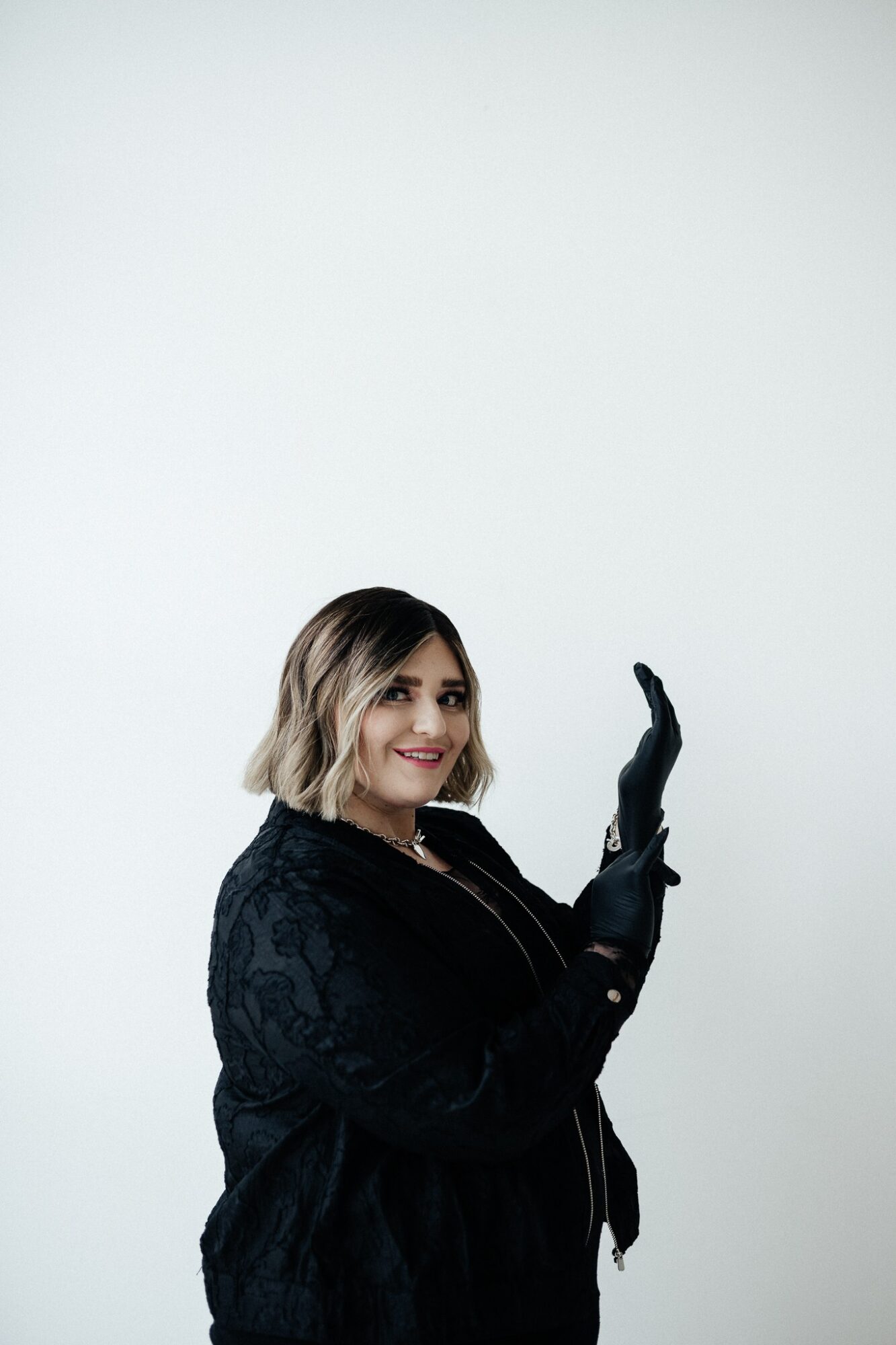 Woman with shoulder-length hair smiling, wearing a dark jacket and gloves, waving with her right hand, against a plain background.