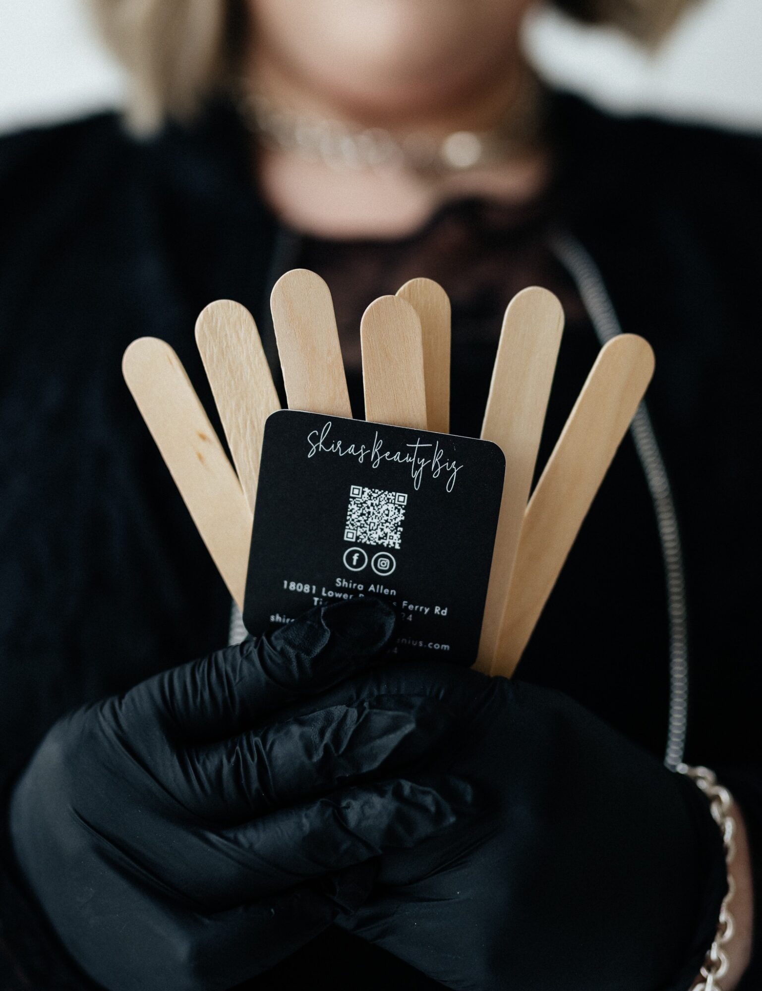 Person wearing black gloves holding a card with a QR code and wooden sticks, with a blurred background.