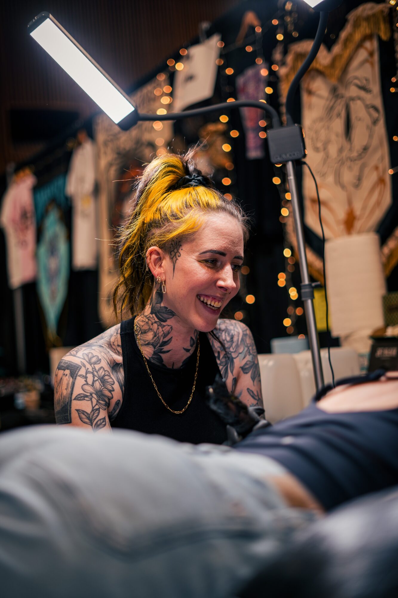 Smiling woman with tattoos and yellow hair in a tattoo studio, surrounded by clothing and warm lights.