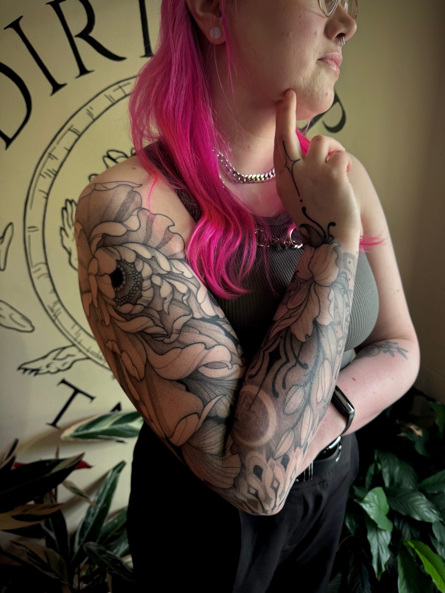 Person with pink hair and tattoos on arm, wearing a necklace, standing near a wall with a circular design, hand on chin.