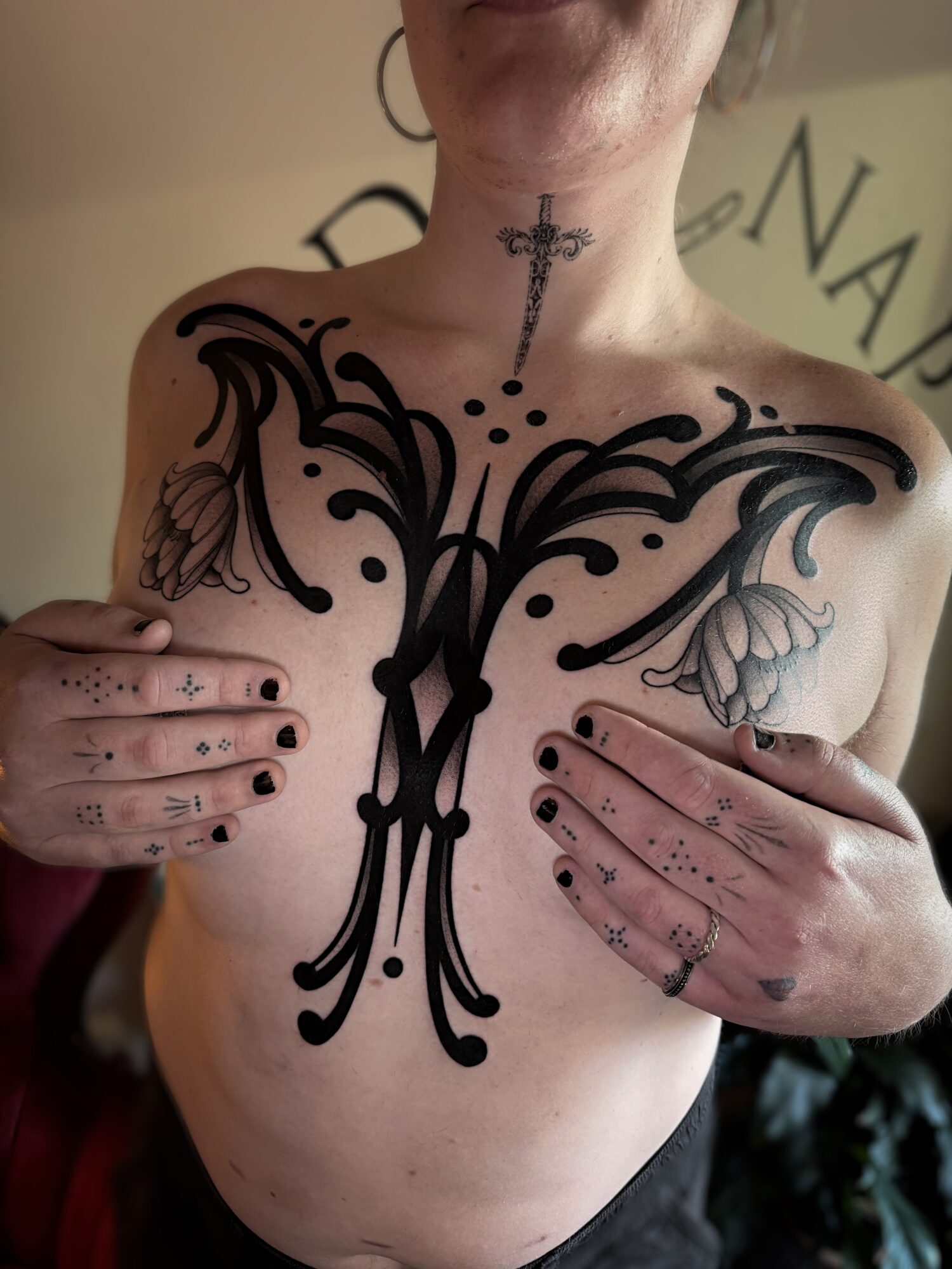 Person with a large black tattoo on chest, holding breasts, with a small cross tattoo on neck, and a ring on finger.