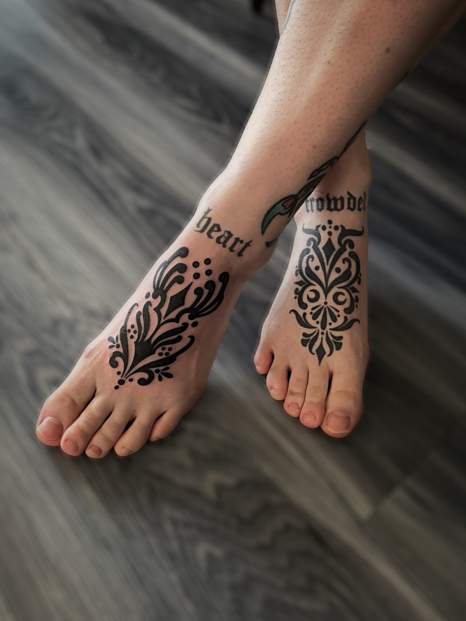 Two feet with black tattoos on ankles and feet, resting on a dark wooden surface.