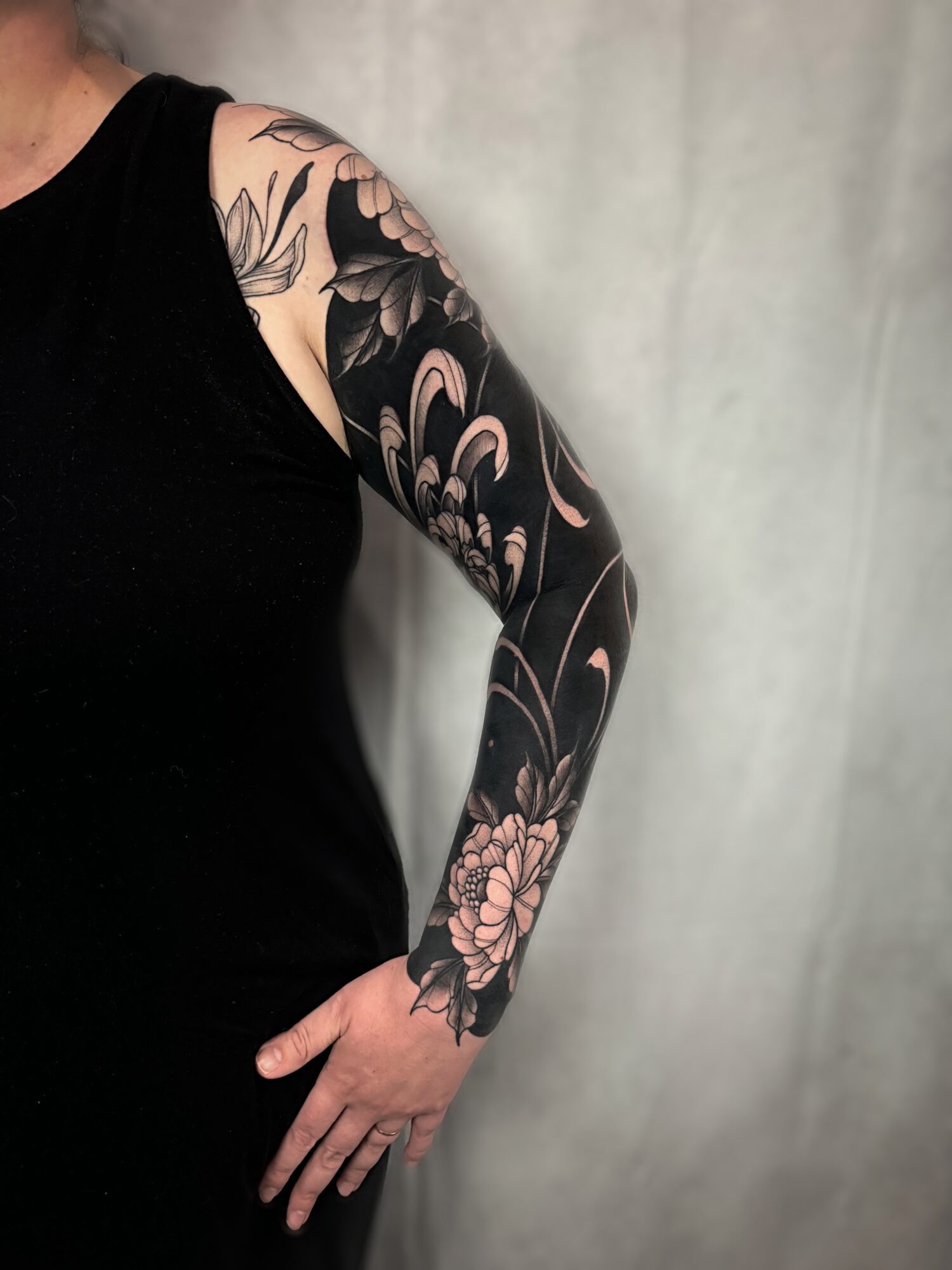 Person with a floral tattoo sleeve on their arm, wearing a black sleeveless top, standing against a plain background.