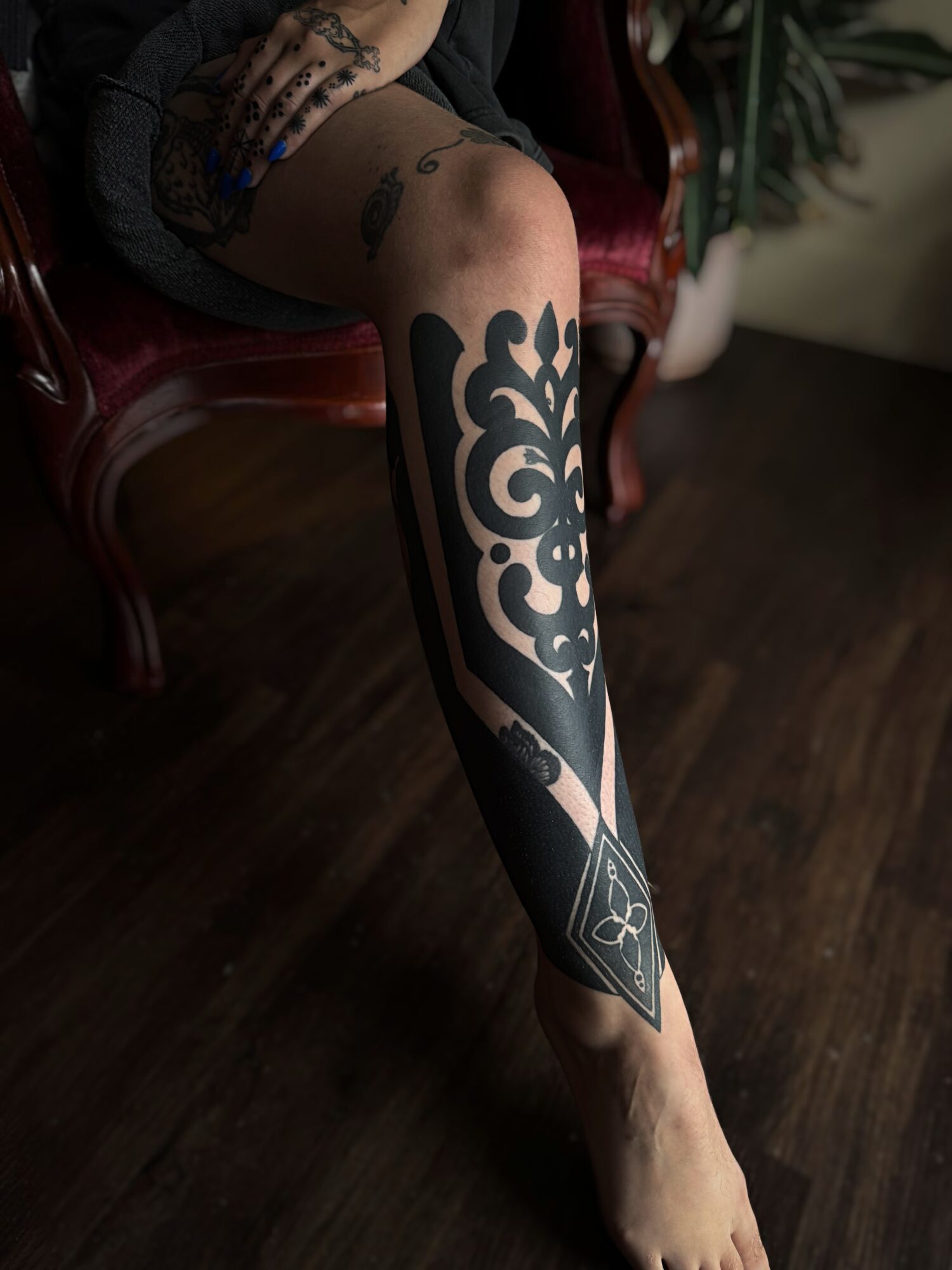 Person sitting on a red chair with a large, black geometric tattoo on their leg, on a dark wooden floor.