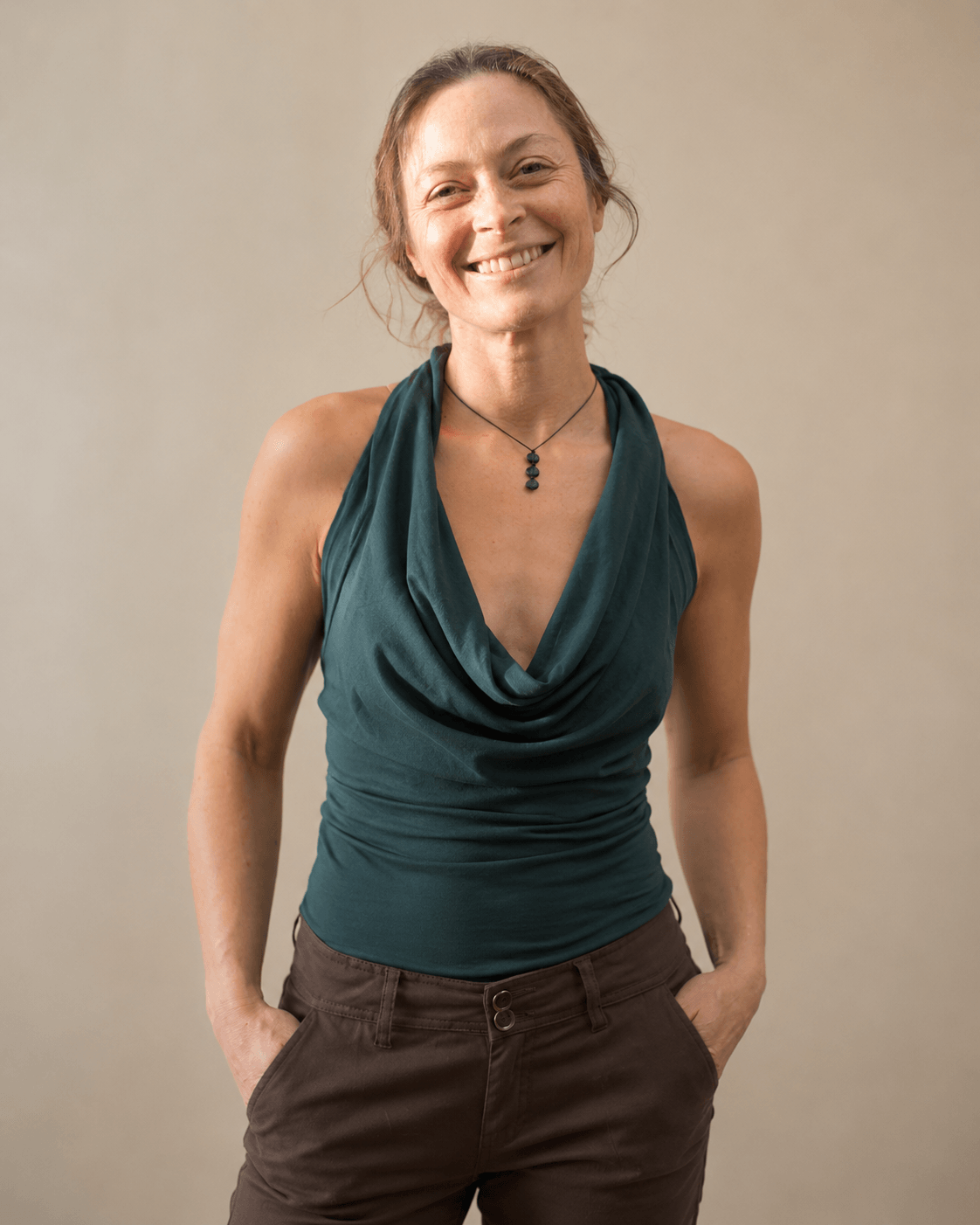 Smiling woman with hands in pockets, wearing a teal sleeveless top and brown pants, standing against a beige background.