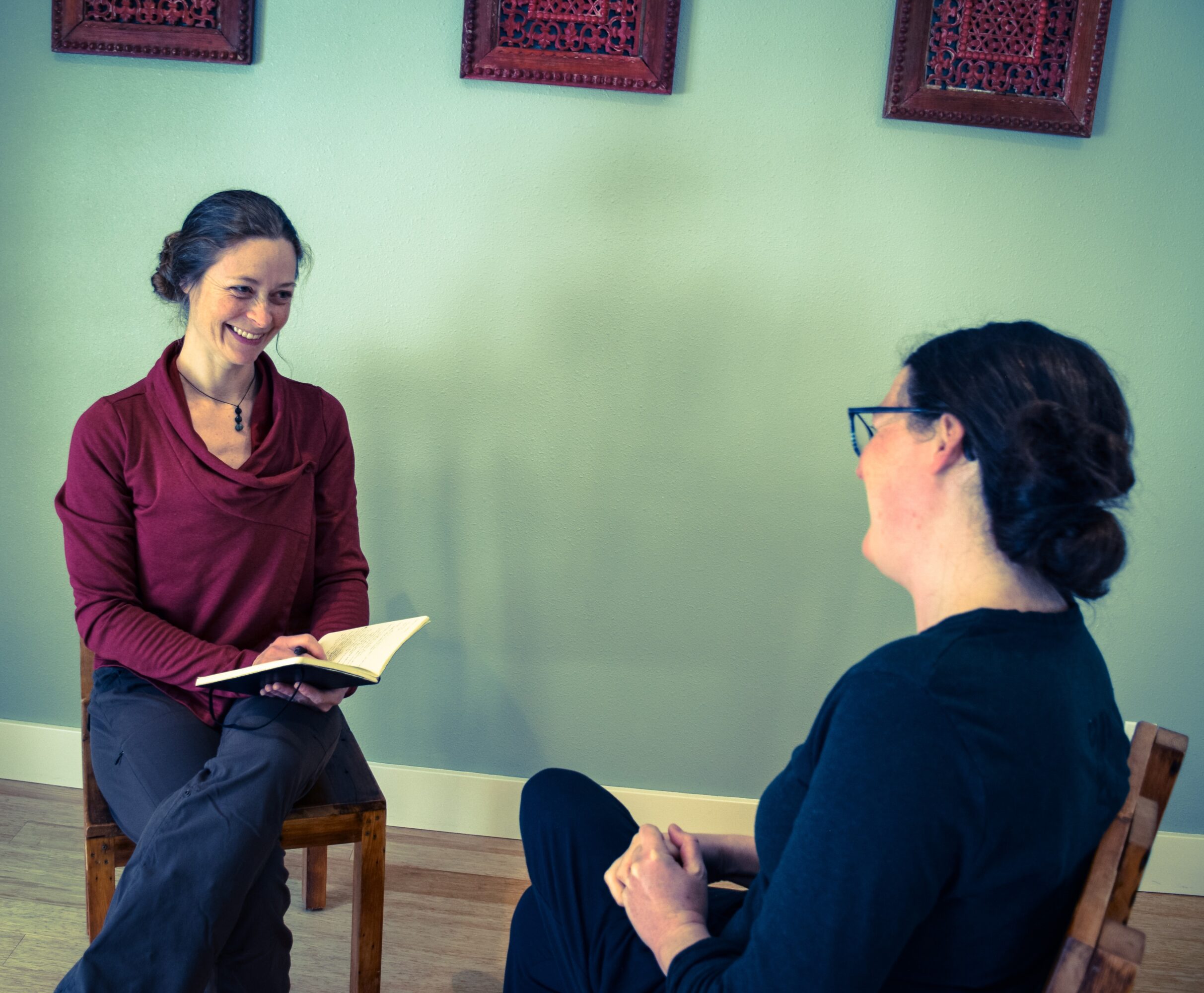 Two women sitting and talking in a room with green walls and framed artwork, one smiling and holding a notebook.