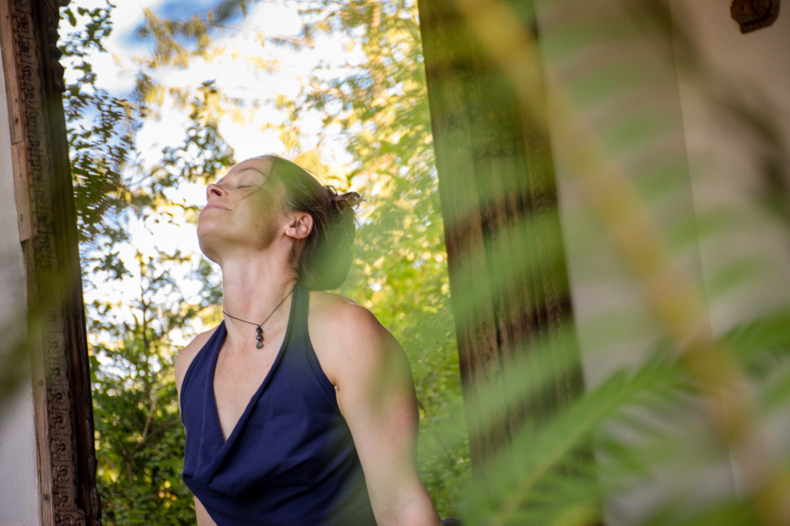 Woman with closed eyes and a smile, standing outdoors among trees and sunlight, wearing a dark blue top.