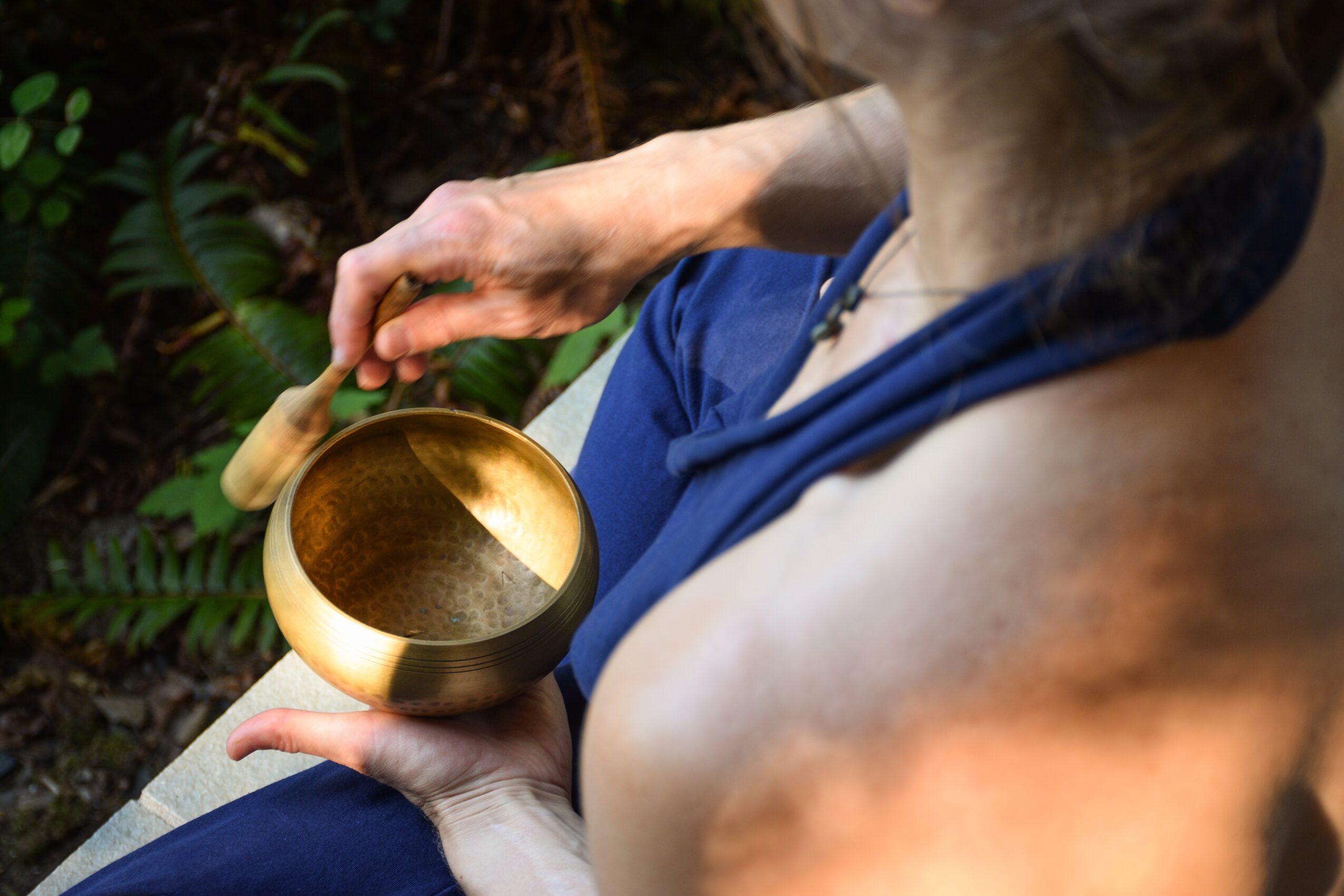 Person holding a golden singing bowl and a wooden mallet outdoors, with greenery in the background.