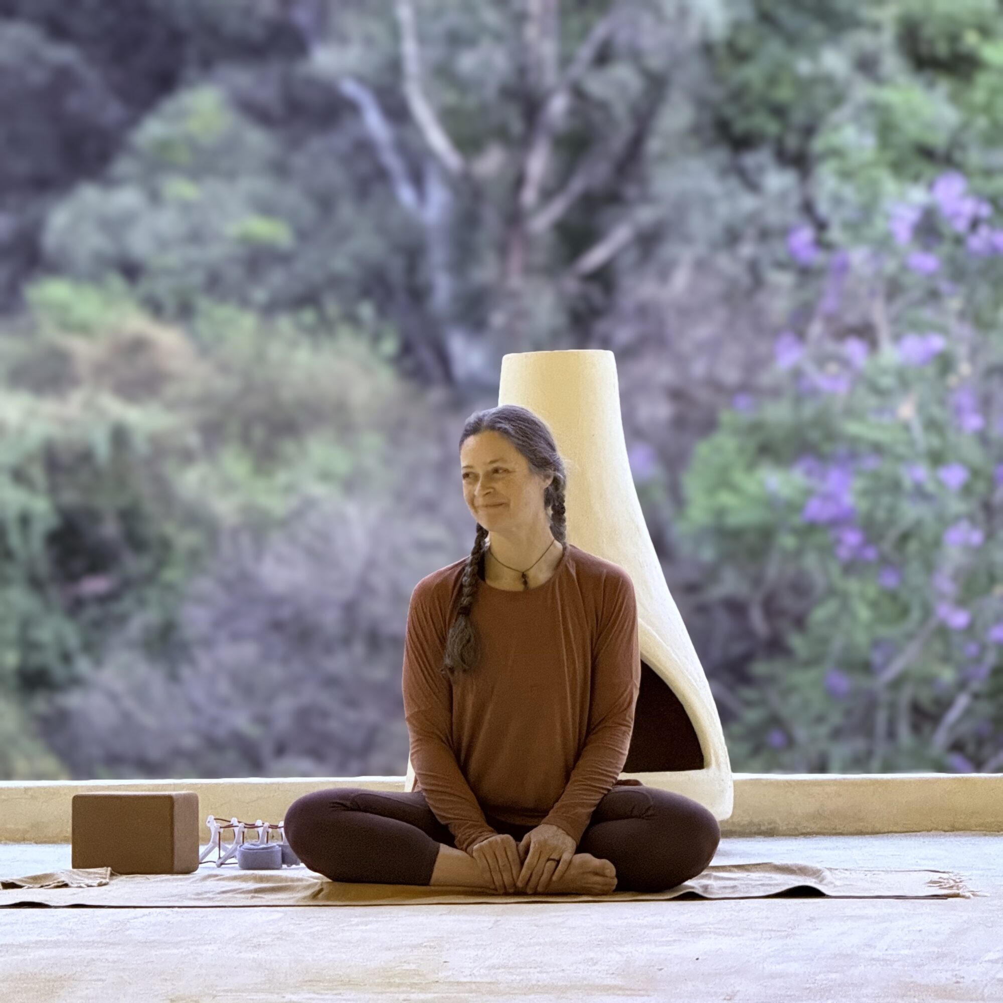 Person sitting cross-legged on a mat indoors with a large window showing trees and greenery outside.