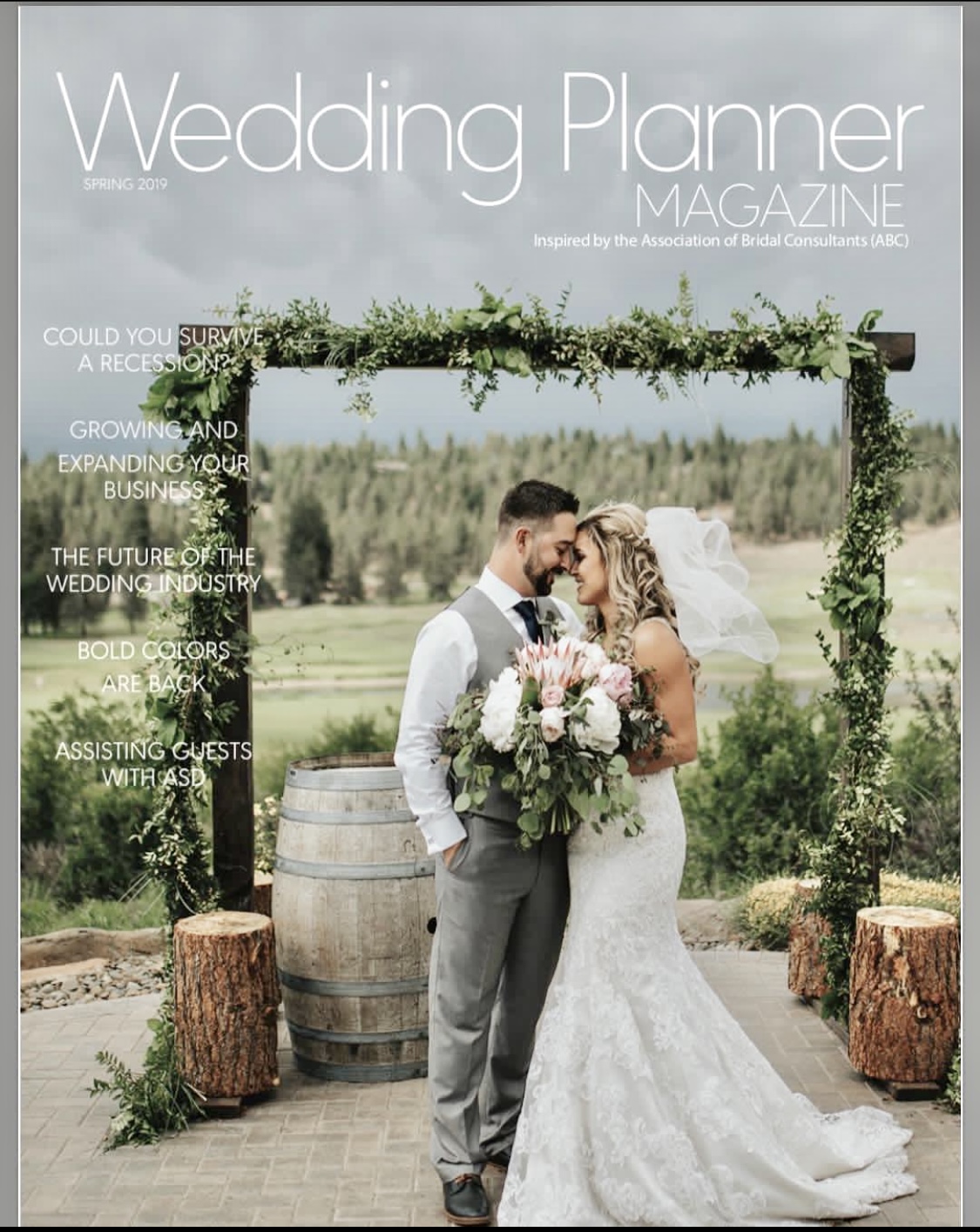 Bride and groom kissing under a decorated arch outdoors, with trees and a field in the background.