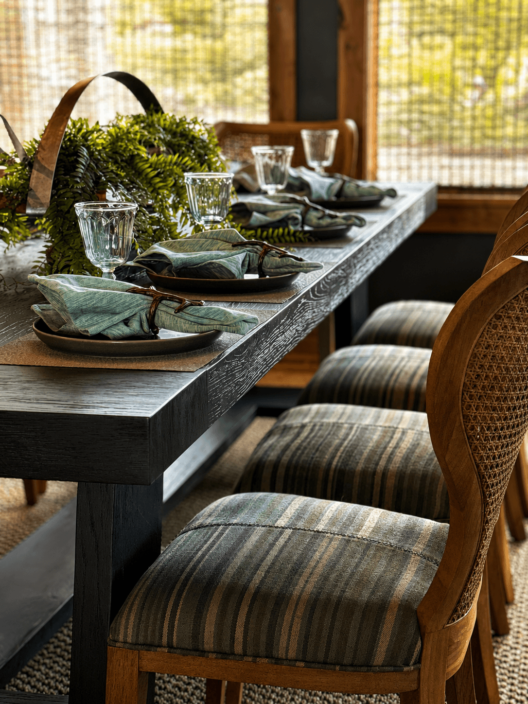 Dining table set with plates, glasses, and napkins, decorated with greenery, near a window with sunlight.