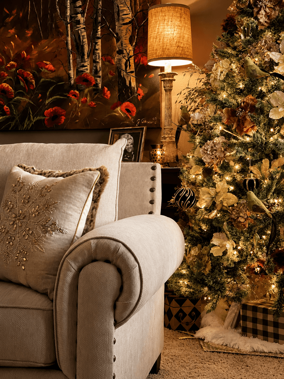 Part of a decorated Christmas tree with ornaments and lights, a sofa with pillows, a lamp, and a floral painting in the background.