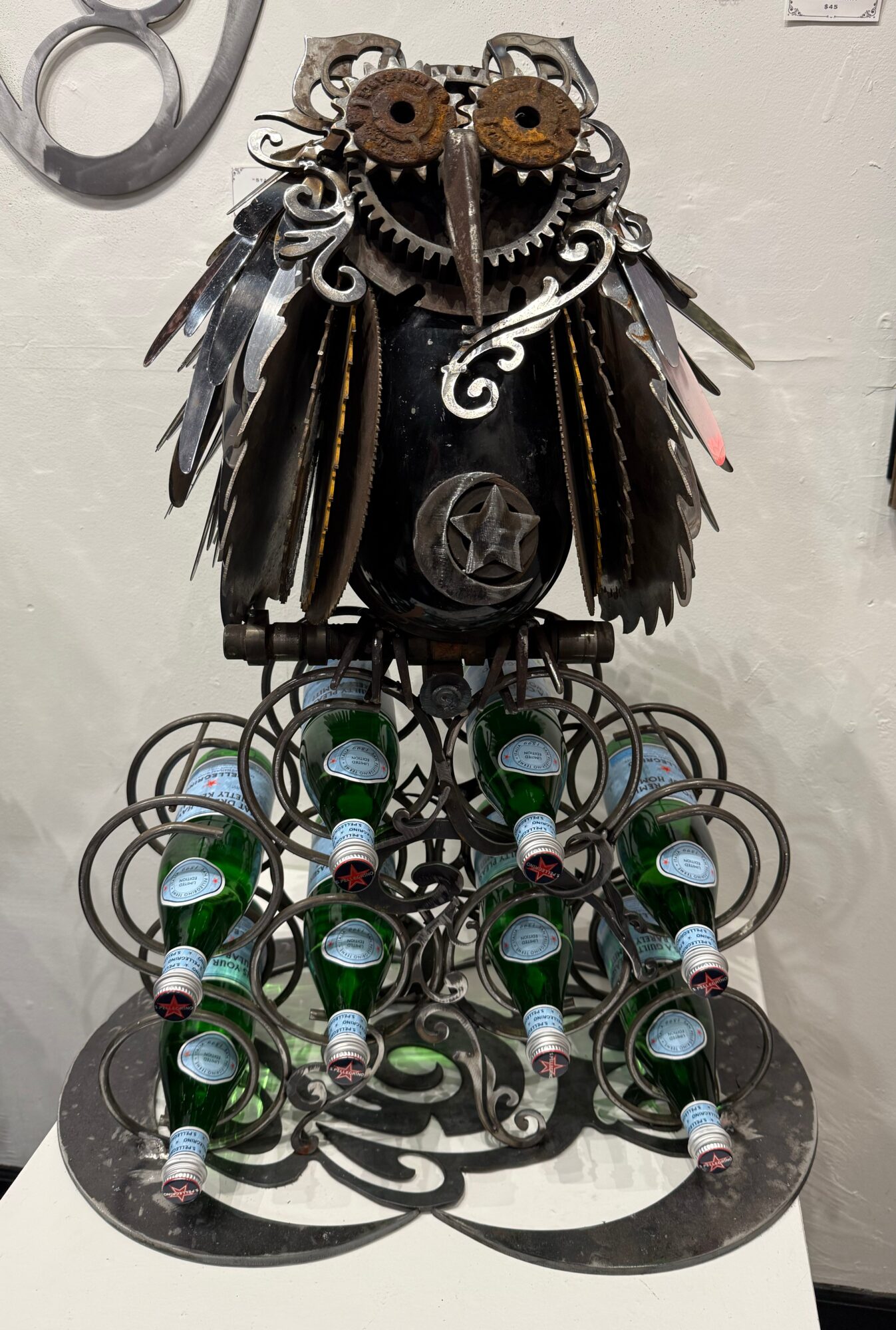 Metal owl sculpture with gears, feathers, and bottles, standing on a decorative base, against a plain wall.