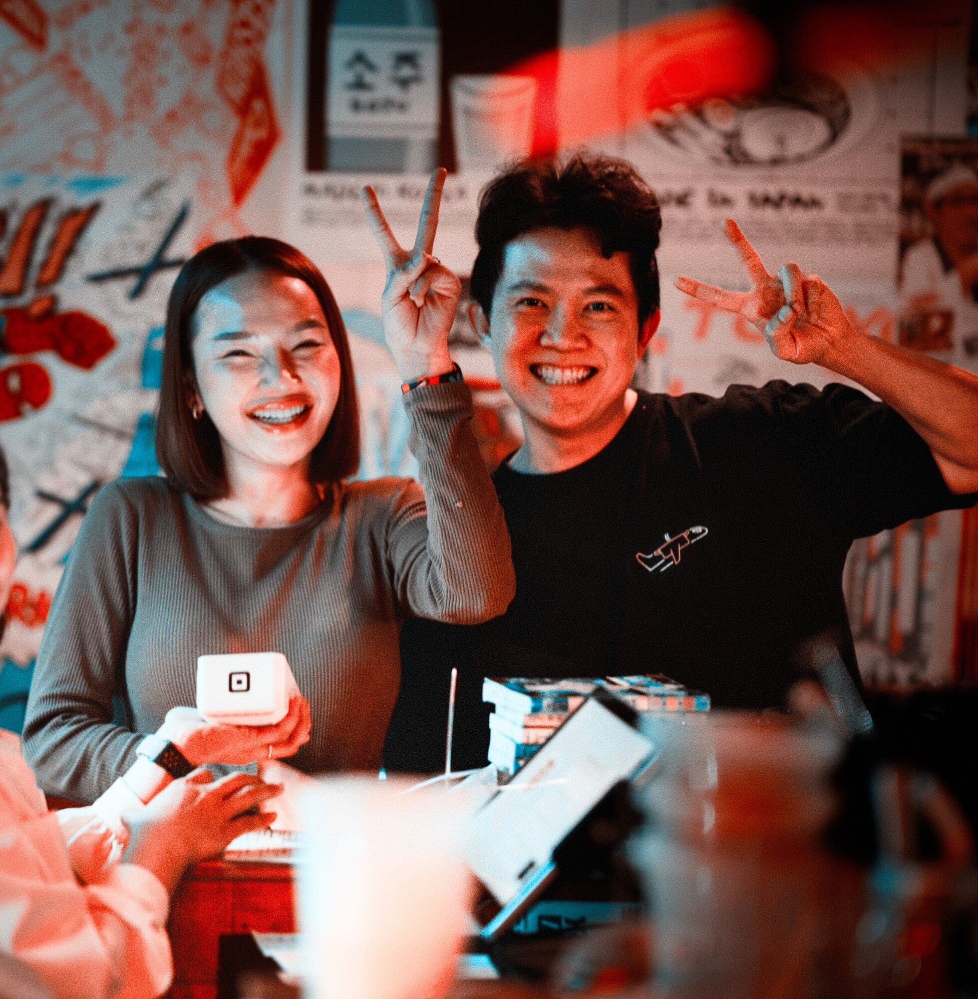 Two smiling people making peace signs, sitting at a table with a blurred background, one holding a phone, in a lively setting.