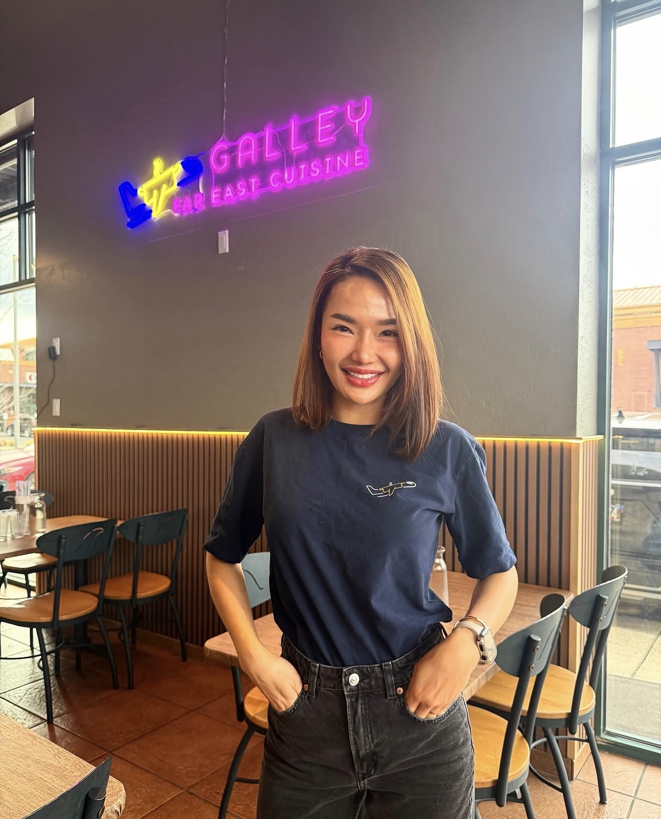 Smiling woman with shoulder-length hair standing inside a cafe, wearing a navy blue t-shirt and black pants, with a neon sign in the background.