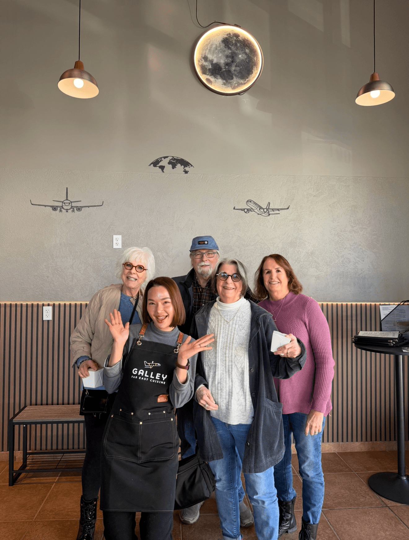Four people stand together in a room with a moon and airplane wall decor, smiling and waving at the camera.