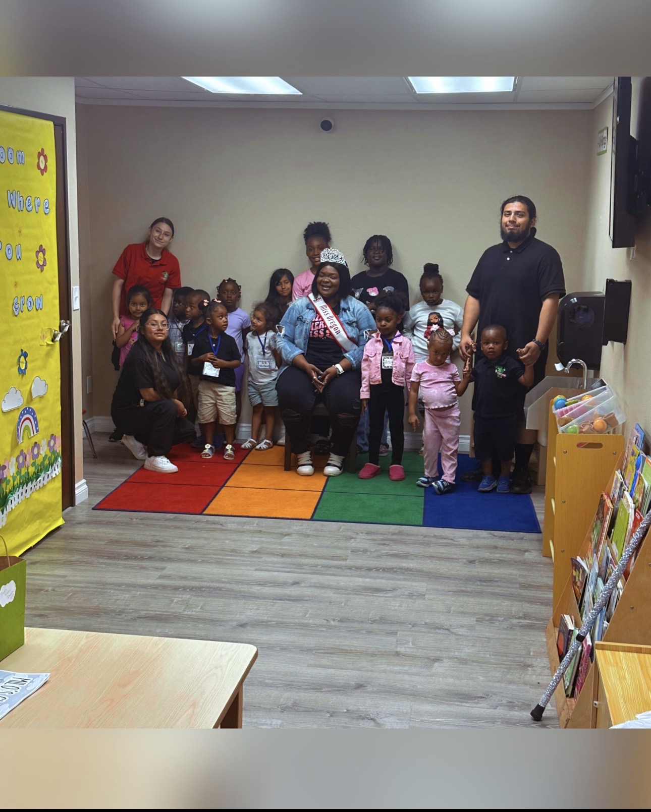 Group of children and adults standing on a colorful mat in a room, with bookshelves and a yellow display on the side.