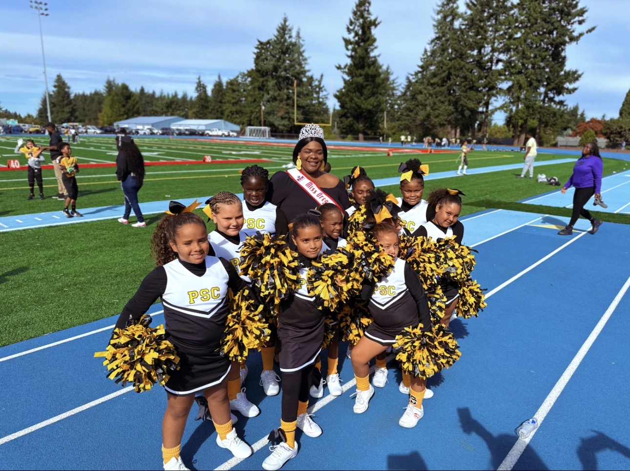Group of young cheerleaders with pom-poms and a coach on a blue track field, with people and trees in background.