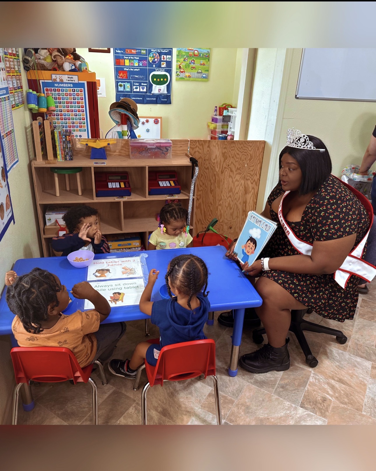 Adult woman reading a book to four children seated at a blue table in a classroom, with educational posters and shelves in the background.
