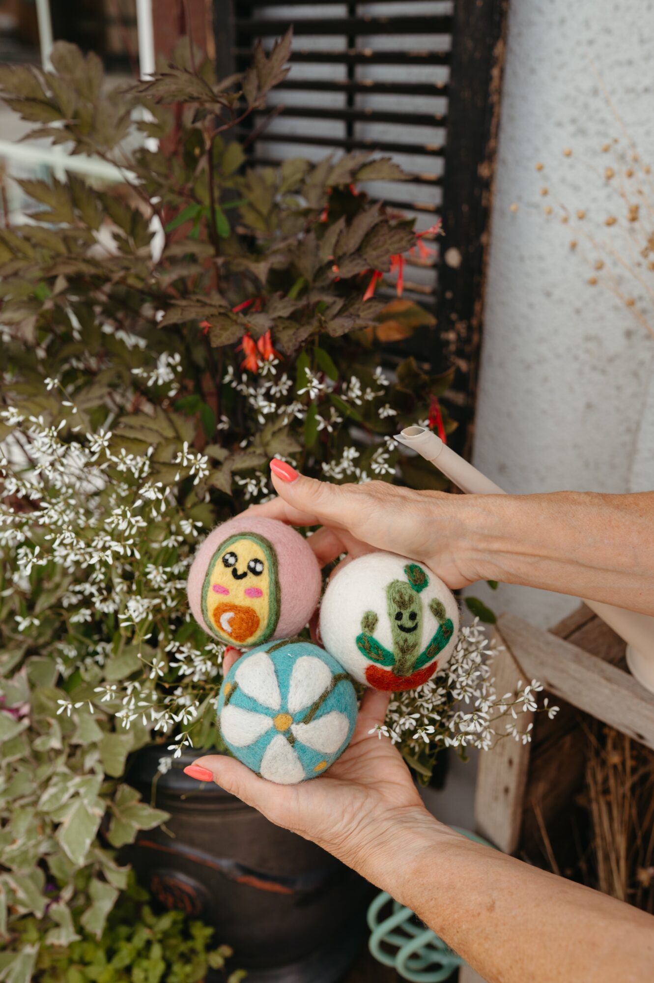 Person holding three decorated eggs with floral and animal designs, outdoors with plants and a fence in background.