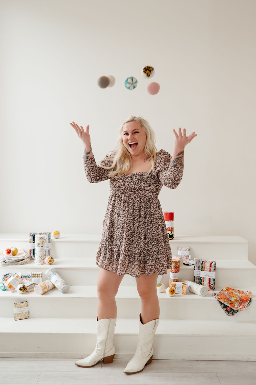 Woman with blonde hair in a floral dress and white boots throws objects in the air, smiling joyfully, against a white background.