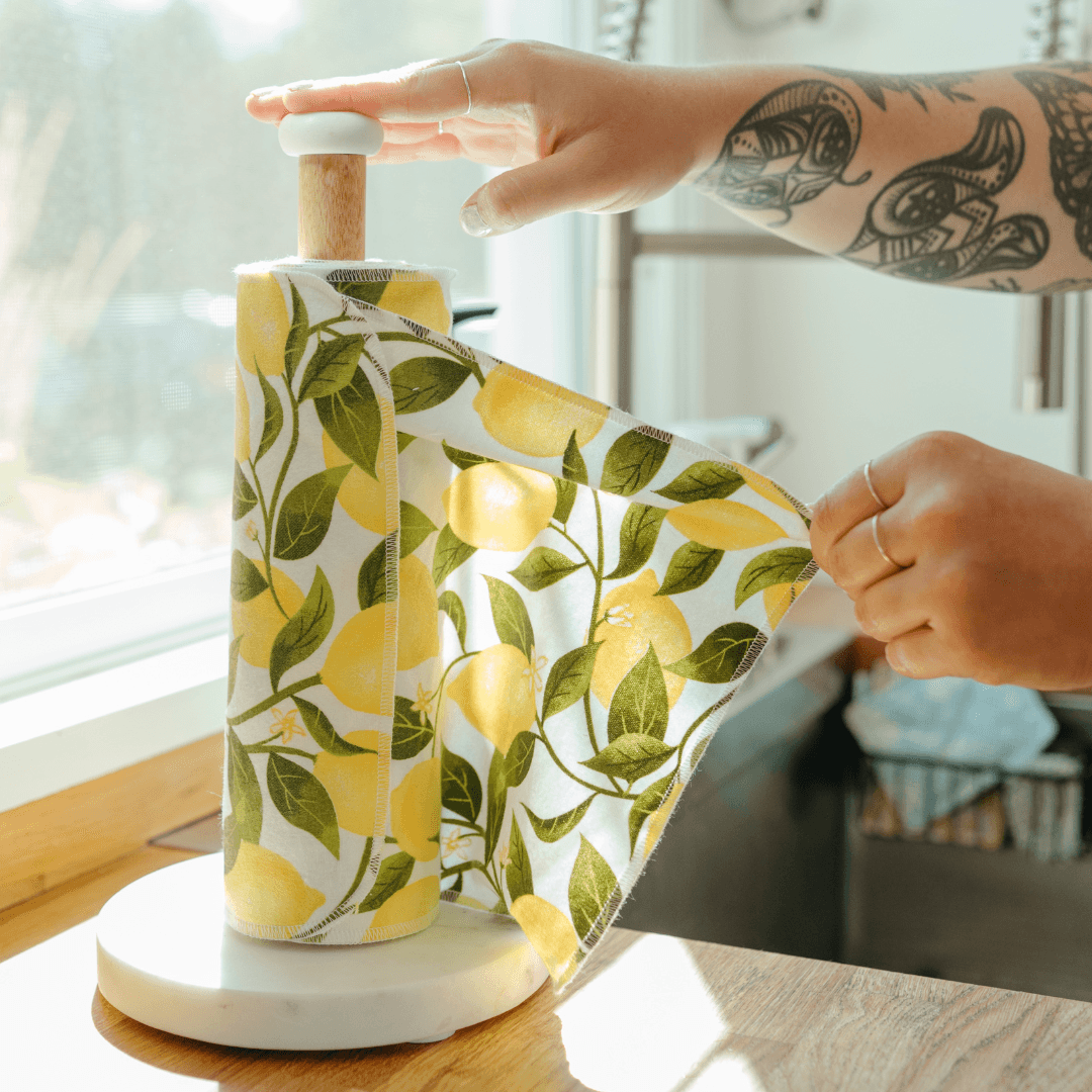 Person with tattooed arm unwrapping lemon-patterned wrapping paper from a bottle on a white base, near a window.