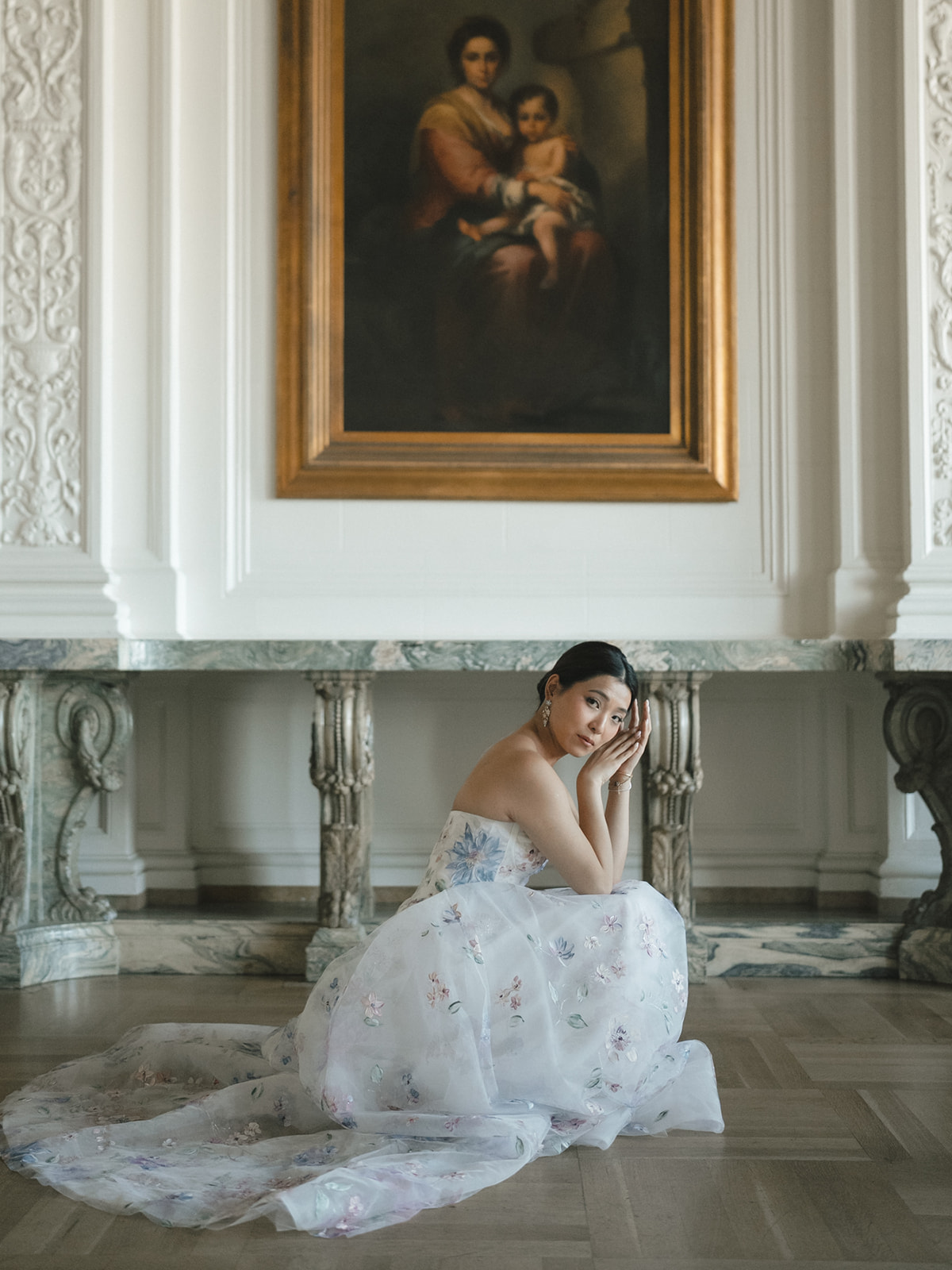 Young woman in a white dress kneels on the floor in front of a large framed painting in an ornate room.