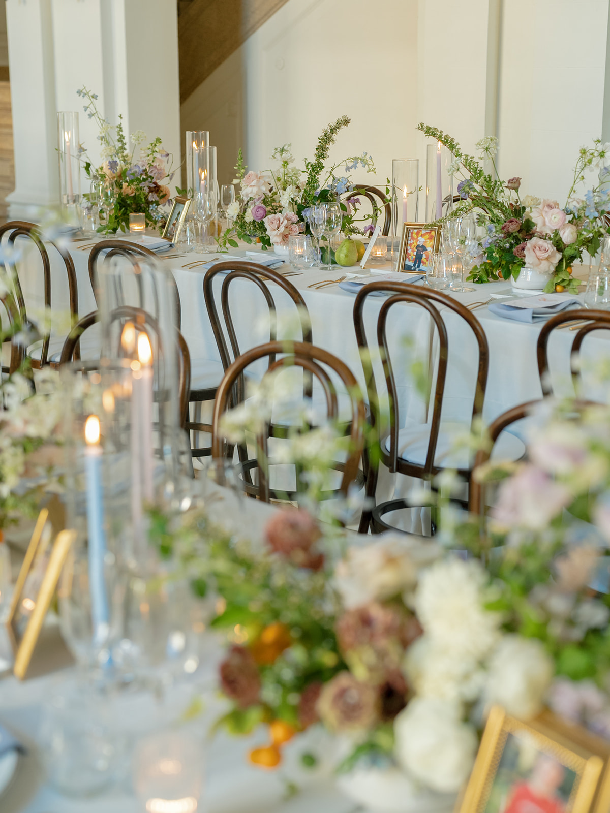 Long table decorated with floral arrangements, candles, and glassware, set for an event in a bright room with white walls.