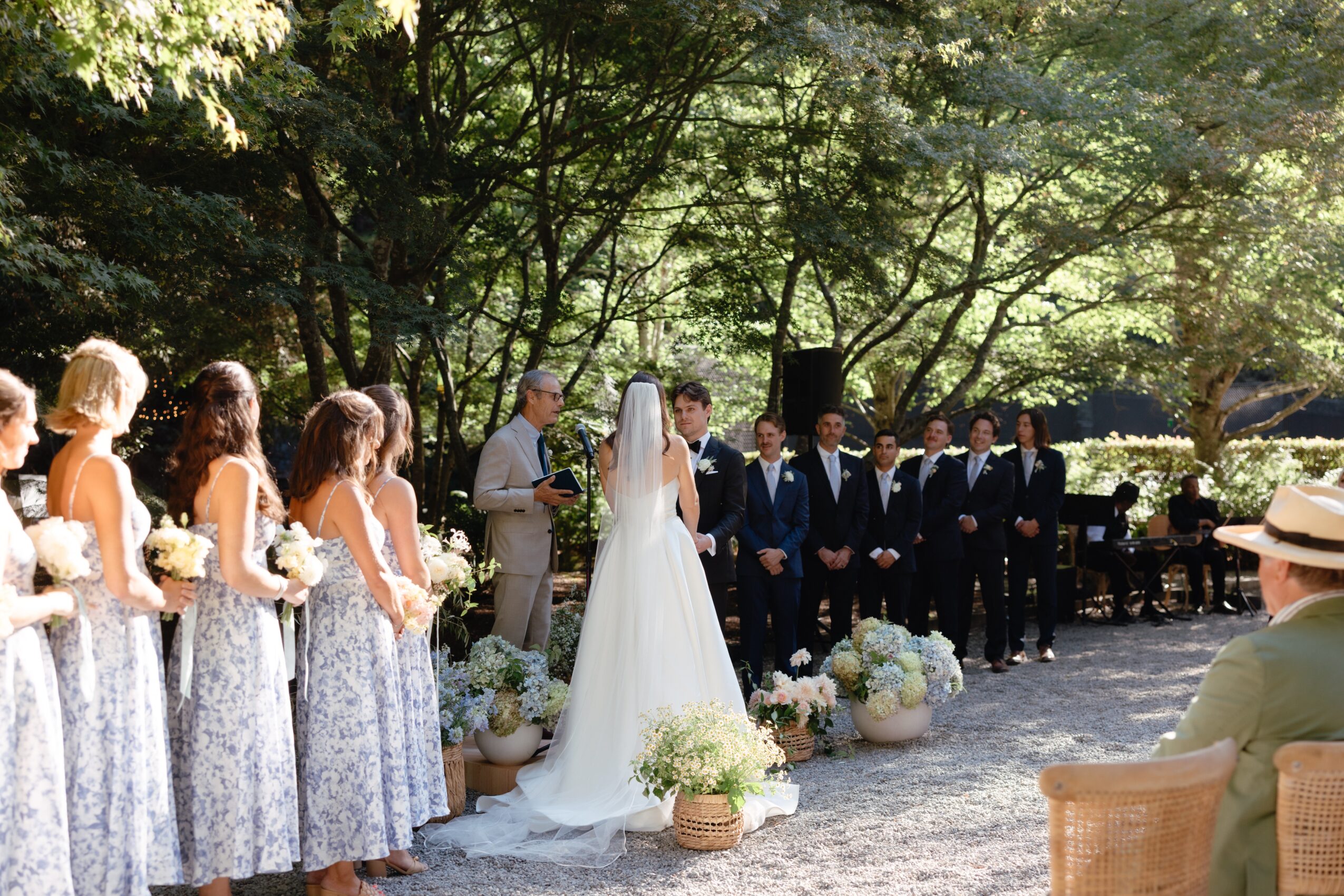 Wedding ceremony outdoors with bride, groom, bridesmaids, and groomsmen standing in a line under trees.