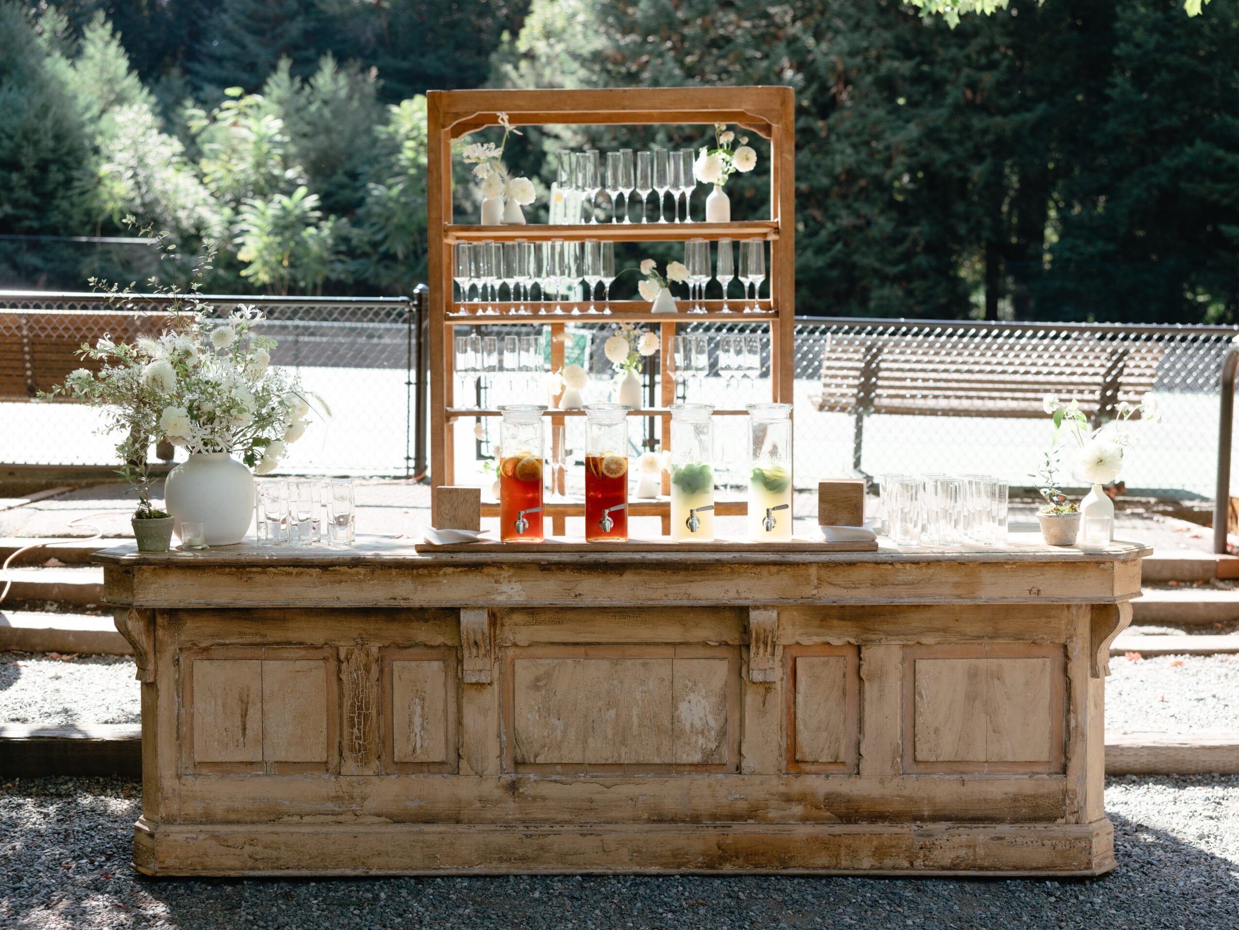 Outdoor bar with glassware and bottles, set against trees and a fence, with sunlight and shadows, rustic wooden counter.