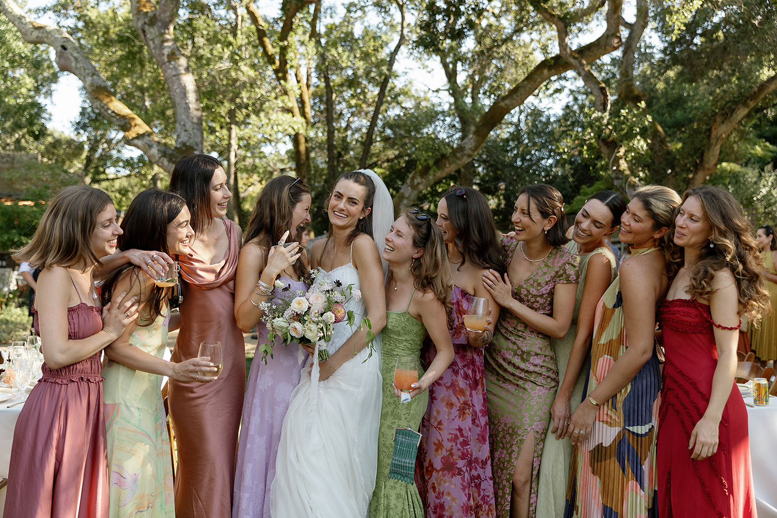 Group of women in colorful dresses gathered outdoors, smiling and talking, with trees and sunlight in the background.