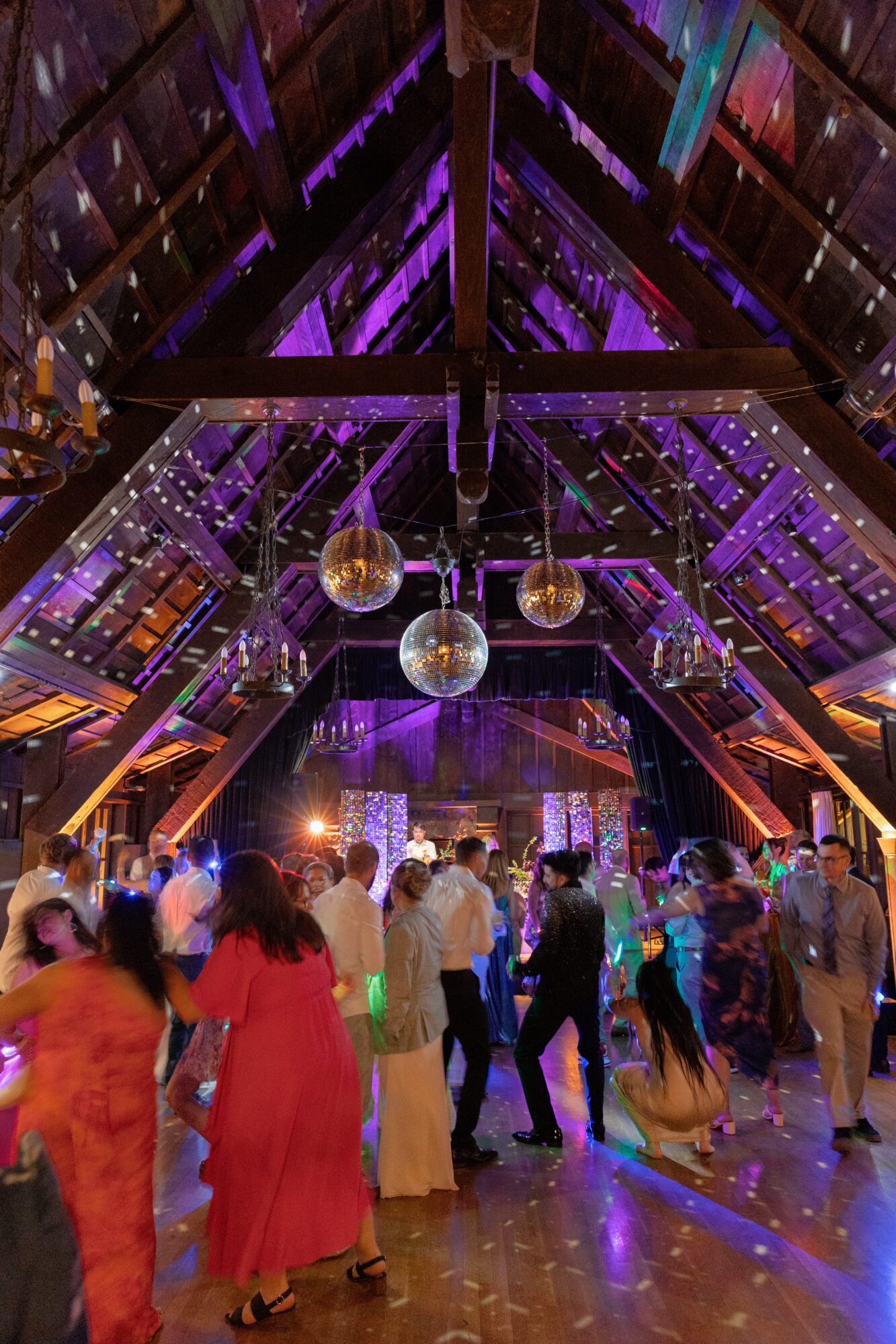 People dancing and socializing in a decorated venue with hanging disco balls and purple lighting, wooden ceiling structure visible.