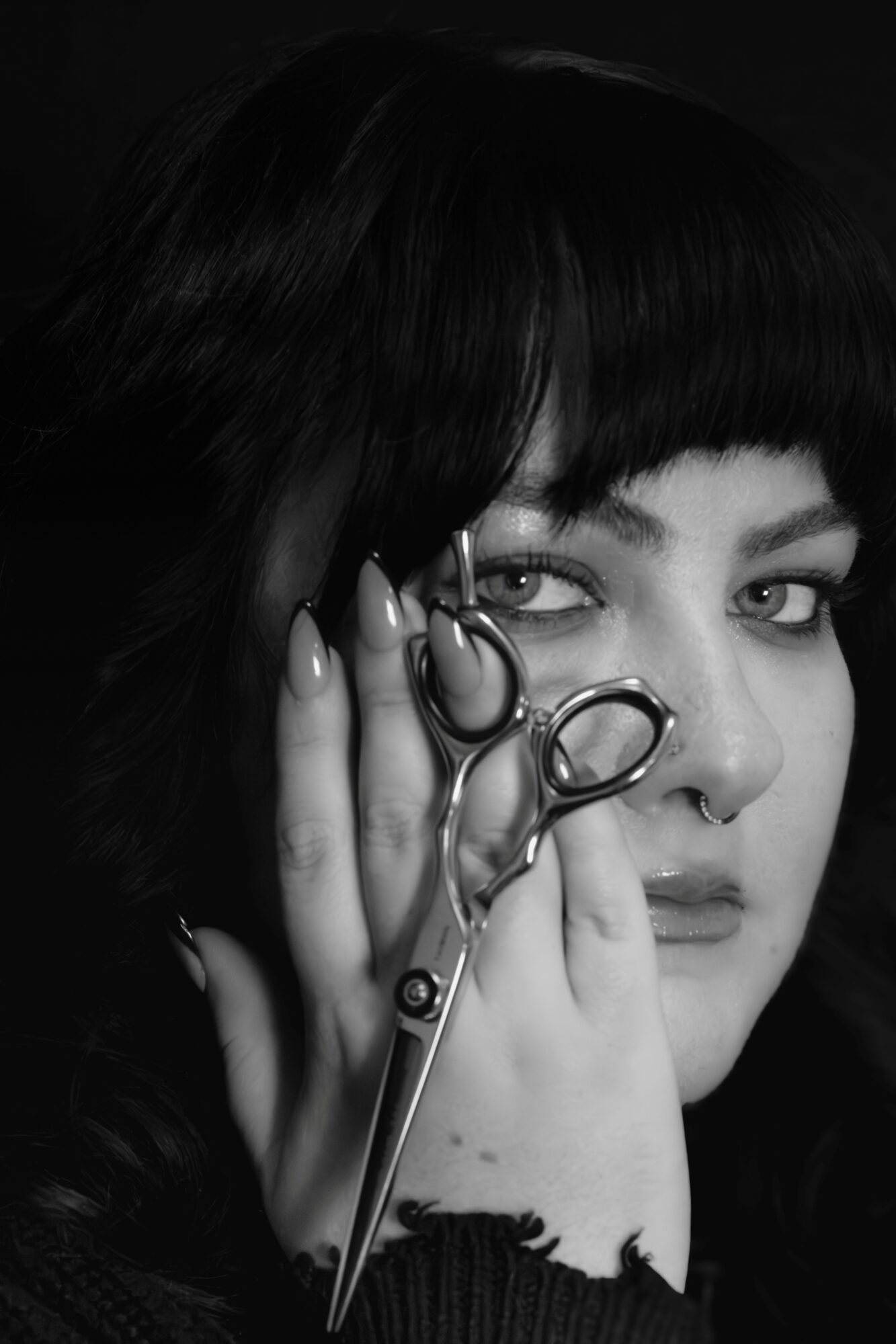 Black and white portrait of a woman with dark hair holding scissors near her face, looking directly at the camera.
