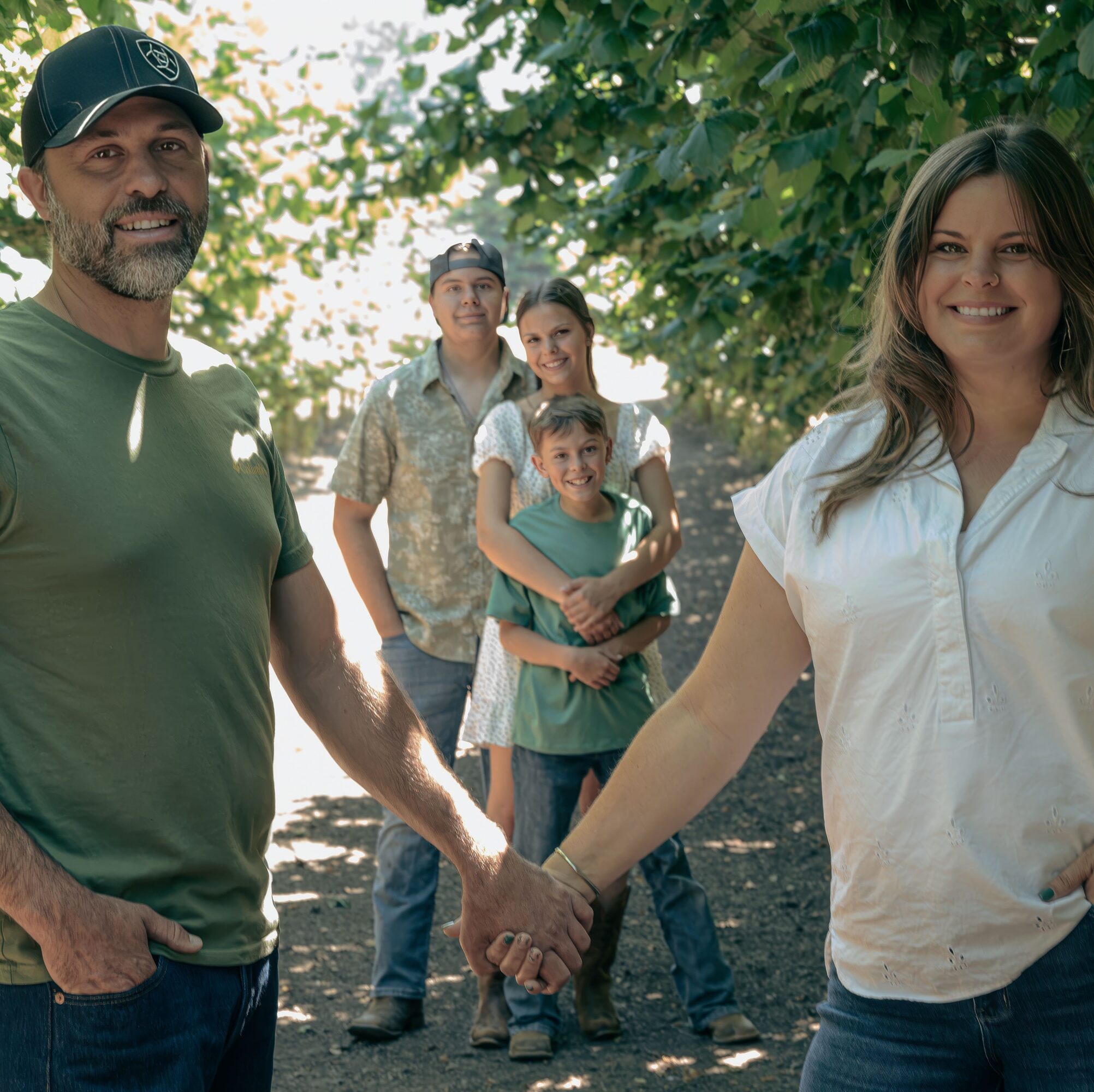 Two adults shake hands in front, three children stand behind them outdoors with trees and sunlight.