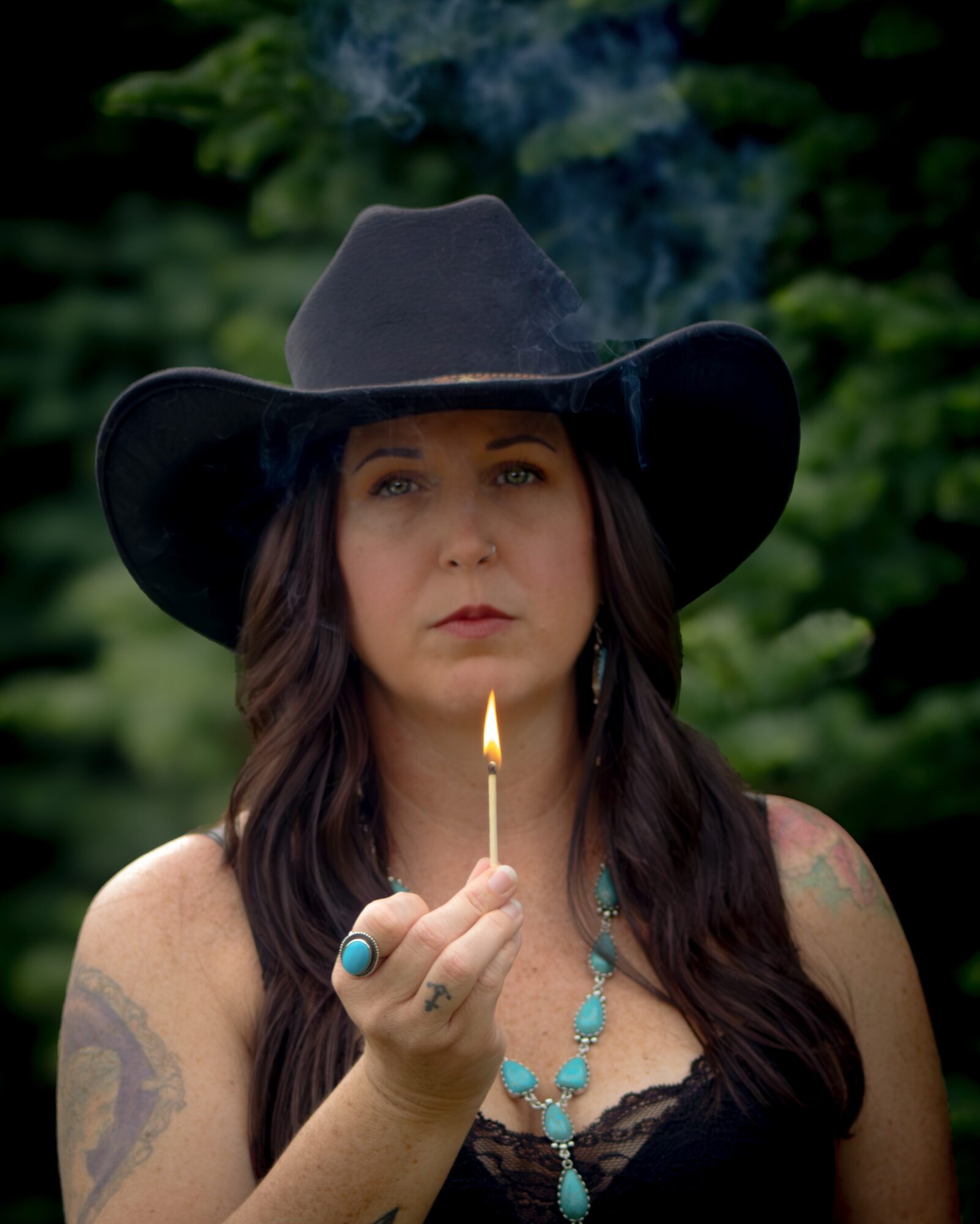 Woman wearing a black hat holding a lit matchstick, outdoors with green foliage background.