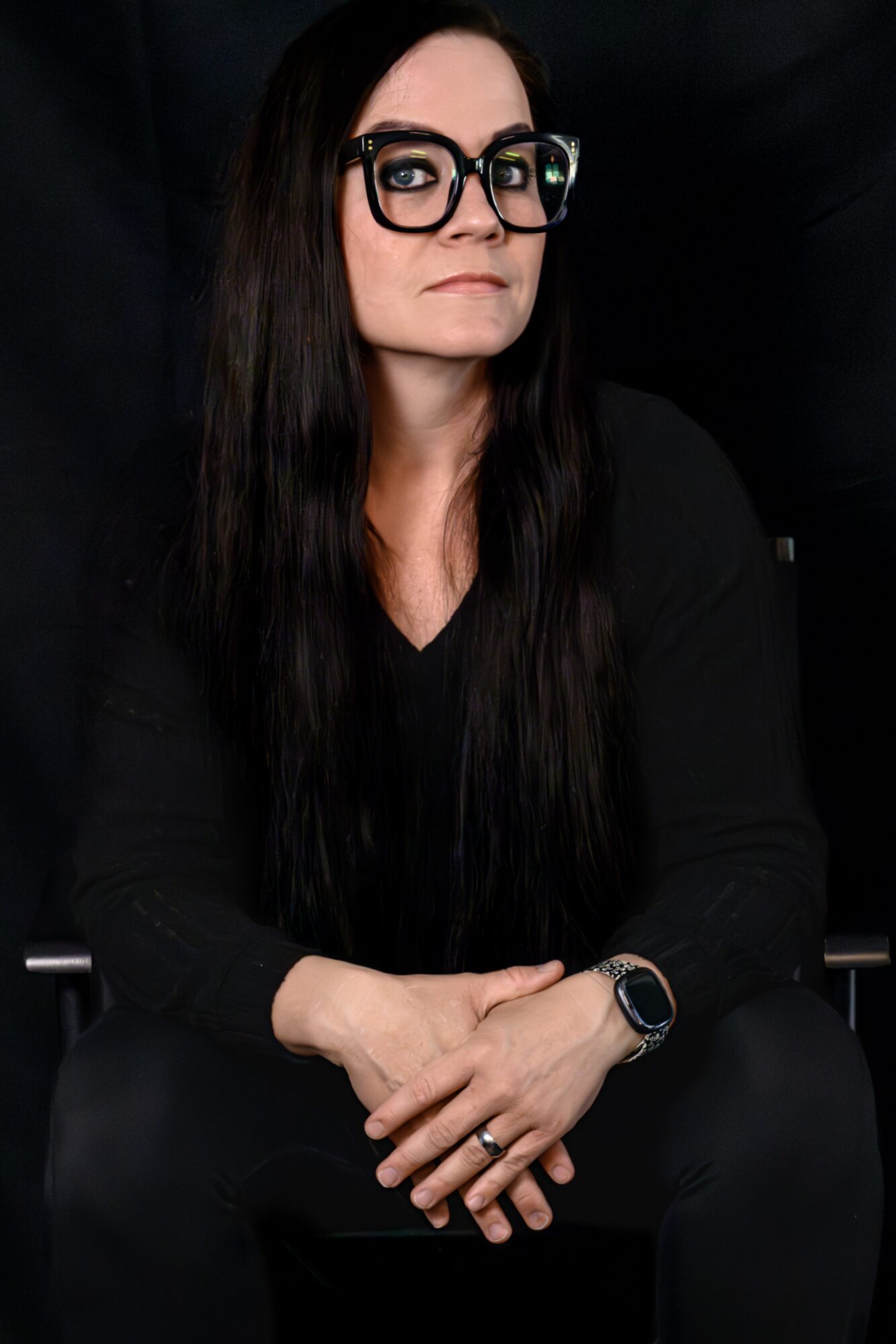 Woman with long dark hair, glasses, and black clothing, sitting with hands crossed, against a dark background.