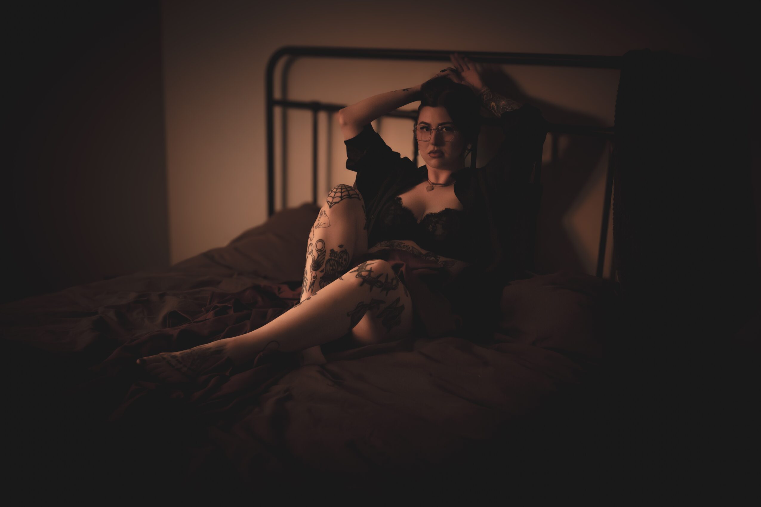 Woman sitting on a bed with her hands in her hair, dimly lit room, dark bedding, relaxed pose.