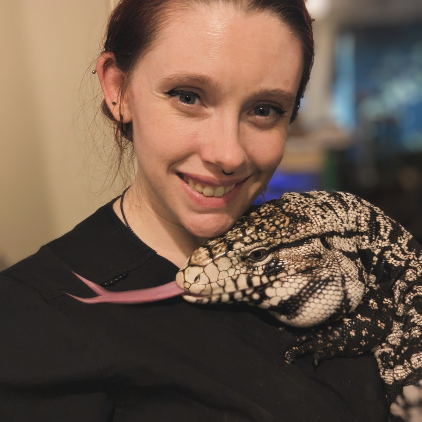 Young woman smiling with a large lizard on her shoulder, indoors, background blurred.