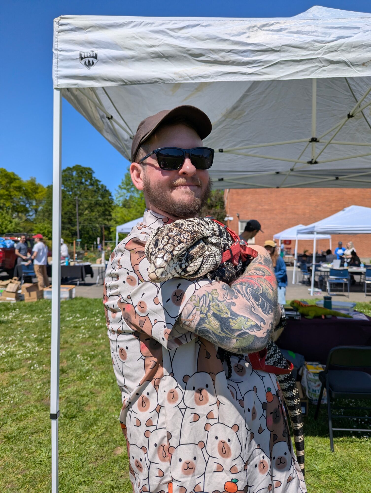 Man with sunglasses and tattoos holding a dog under a white canopy outdoors, with people and buildings in background.