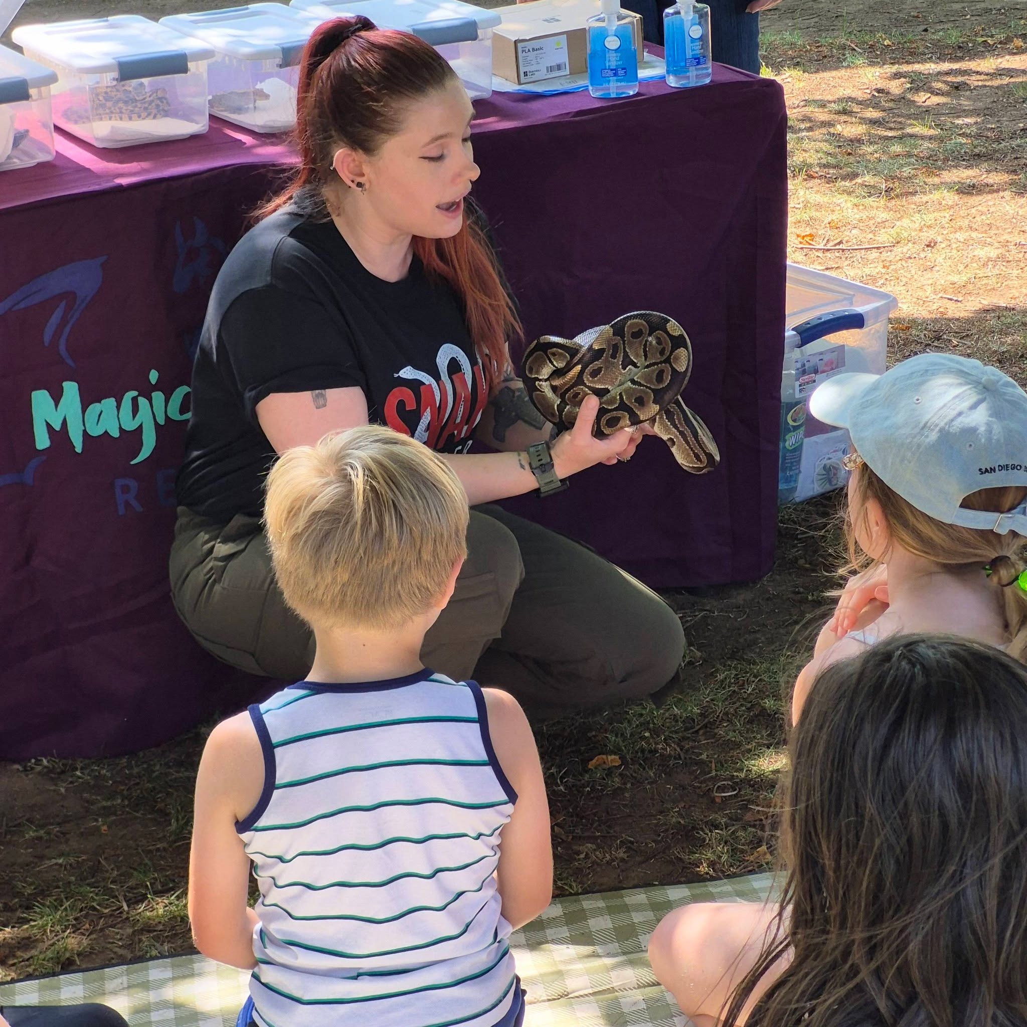 A woman shows a snake to children outdoors at a table with containers and supplies, during daytime.