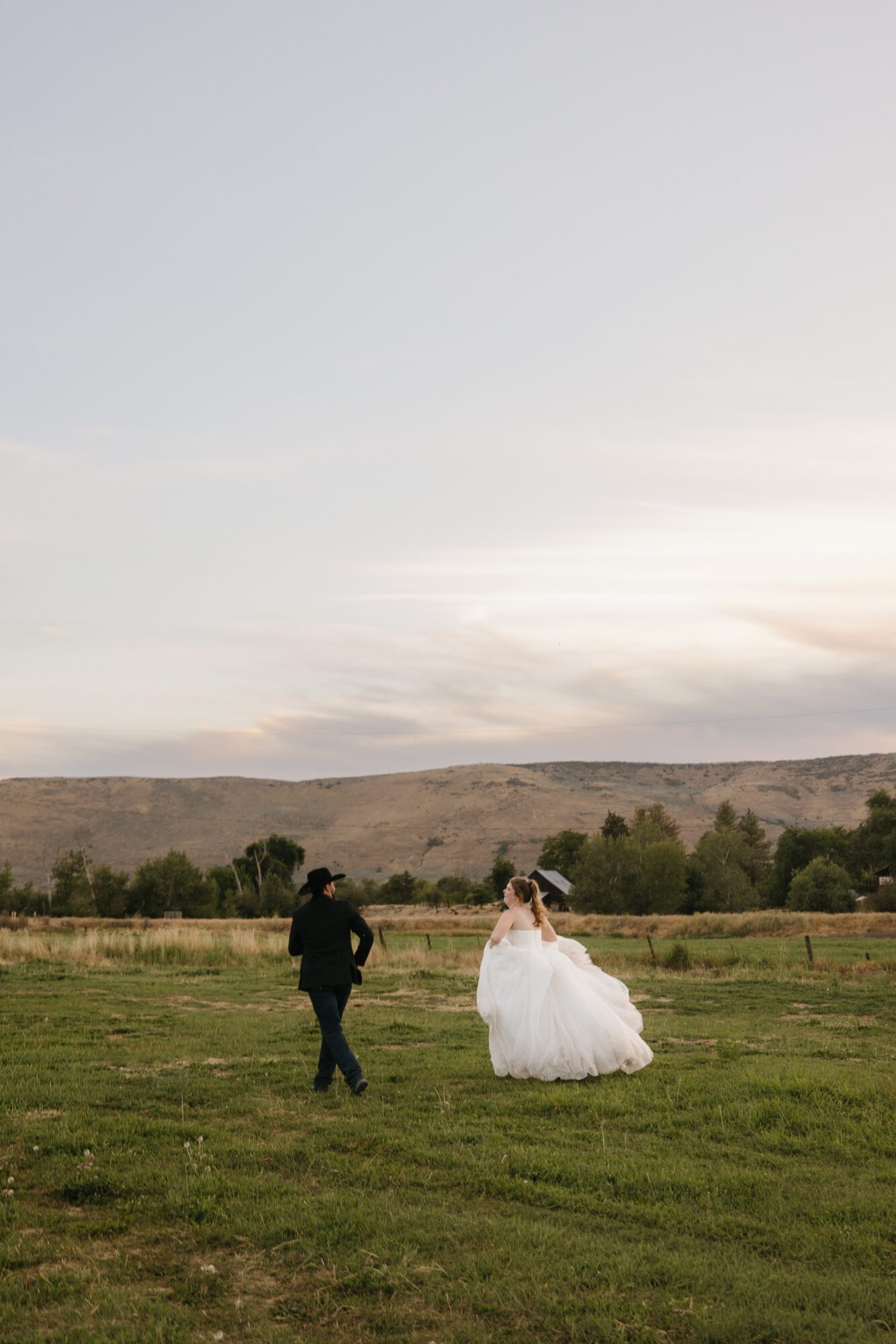 A couple walking in a grassy field with a distant landscape and cloudy sky, one in a wedding dress and the other in dark clothing.