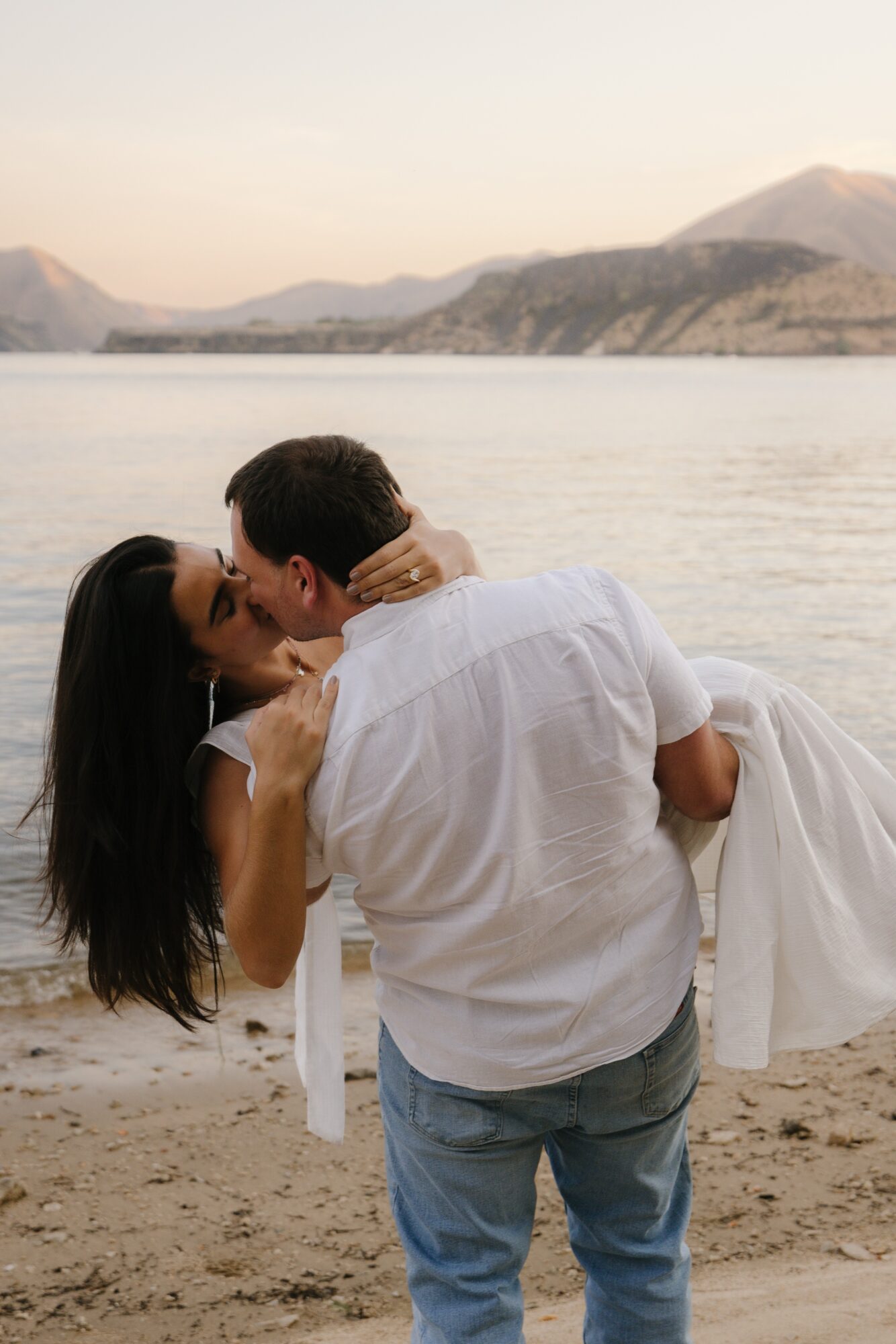 A couple embraces on a beach with mountains and water in the background, the woman is being lifted by the man.