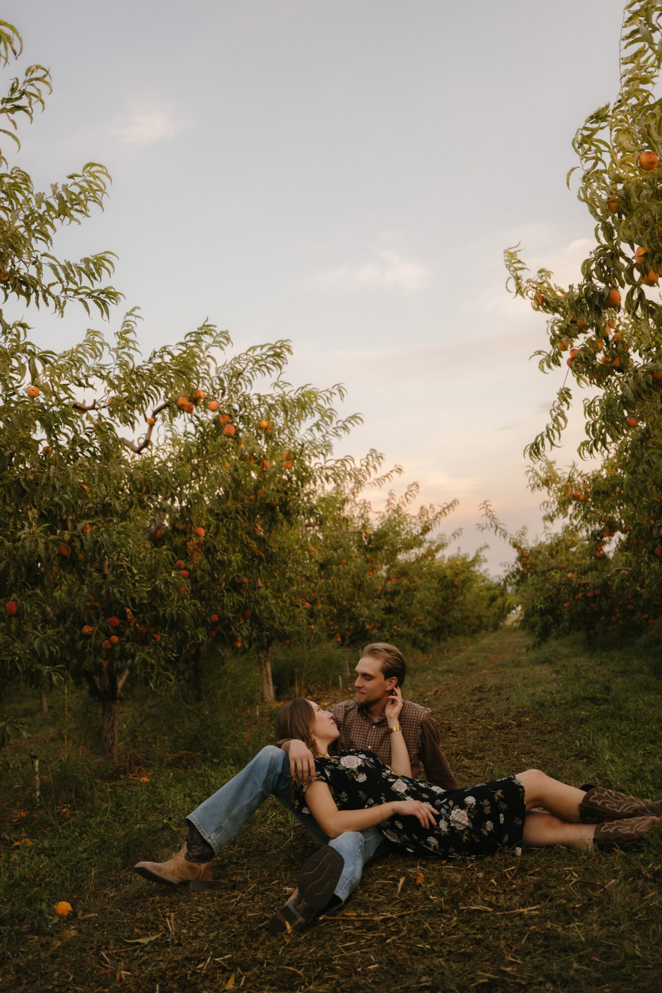 Two people sit on the ground in an orchard with trees and fruit, under a clear sky, embracing each other.