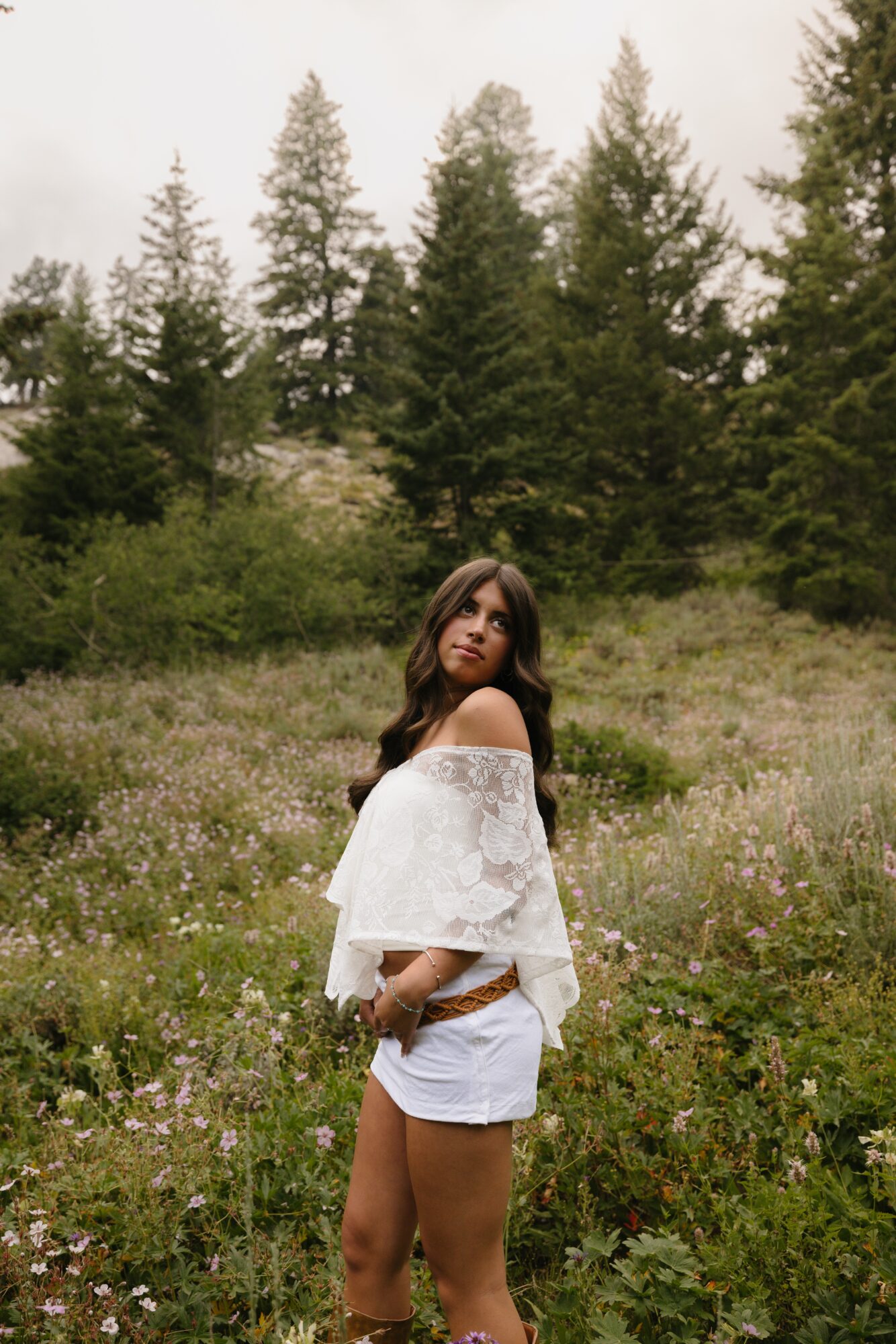 Young woman standing in a field with trees in the background, wearing a white off-shoulder top and shorts.