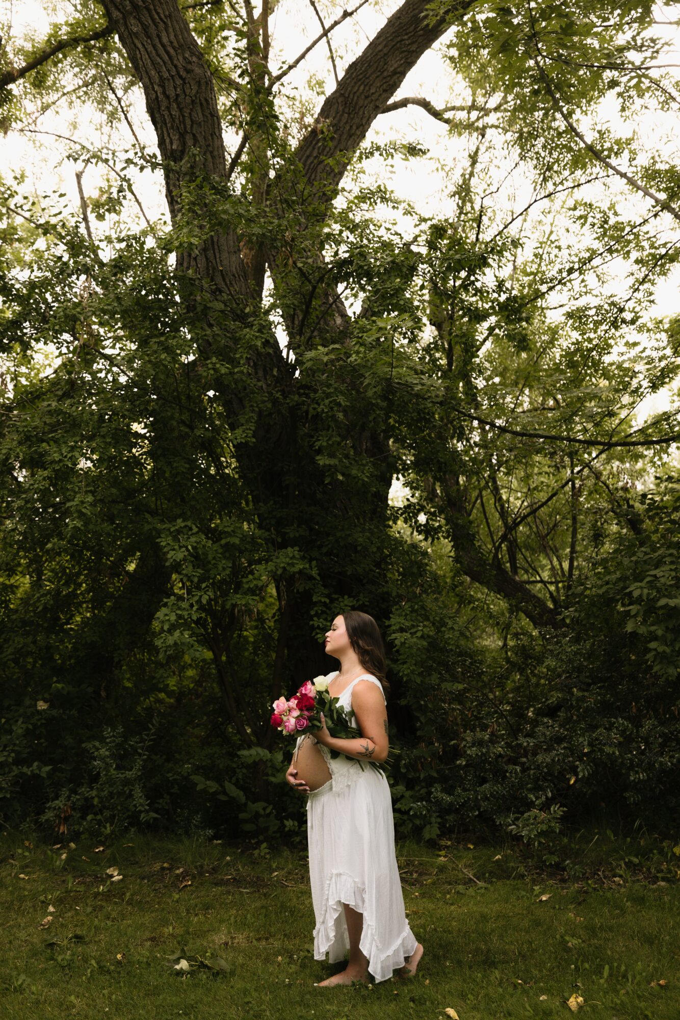 Woman in white dress holding a bouquet of flowers standing outdoors near large trees and greenery